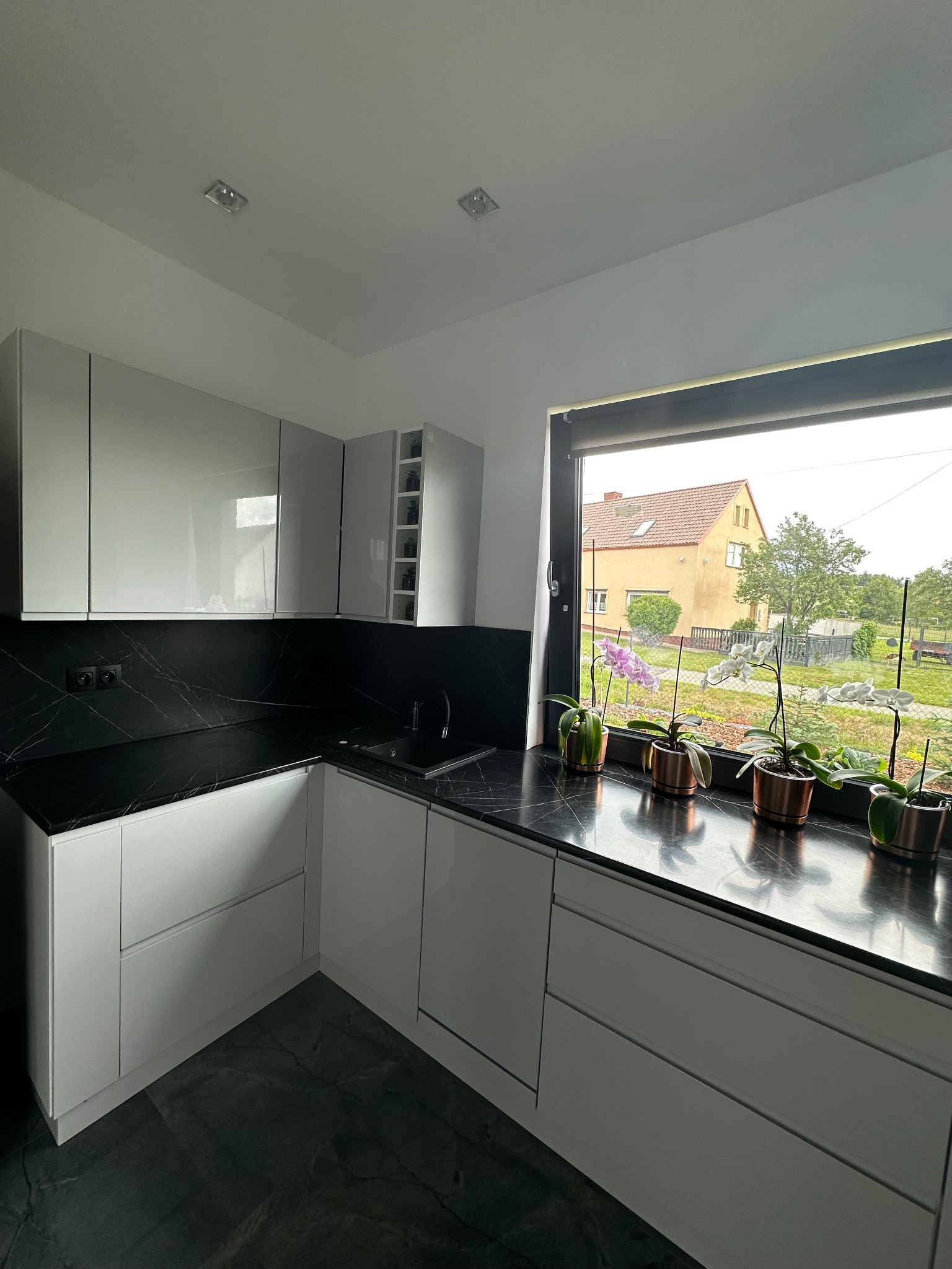 Nowoczesna, narożna, biała kuchnia z czarnym blatem i backsplashiem imitującym marmur, widok na ogród przez okno z doniczkami z orchideami.