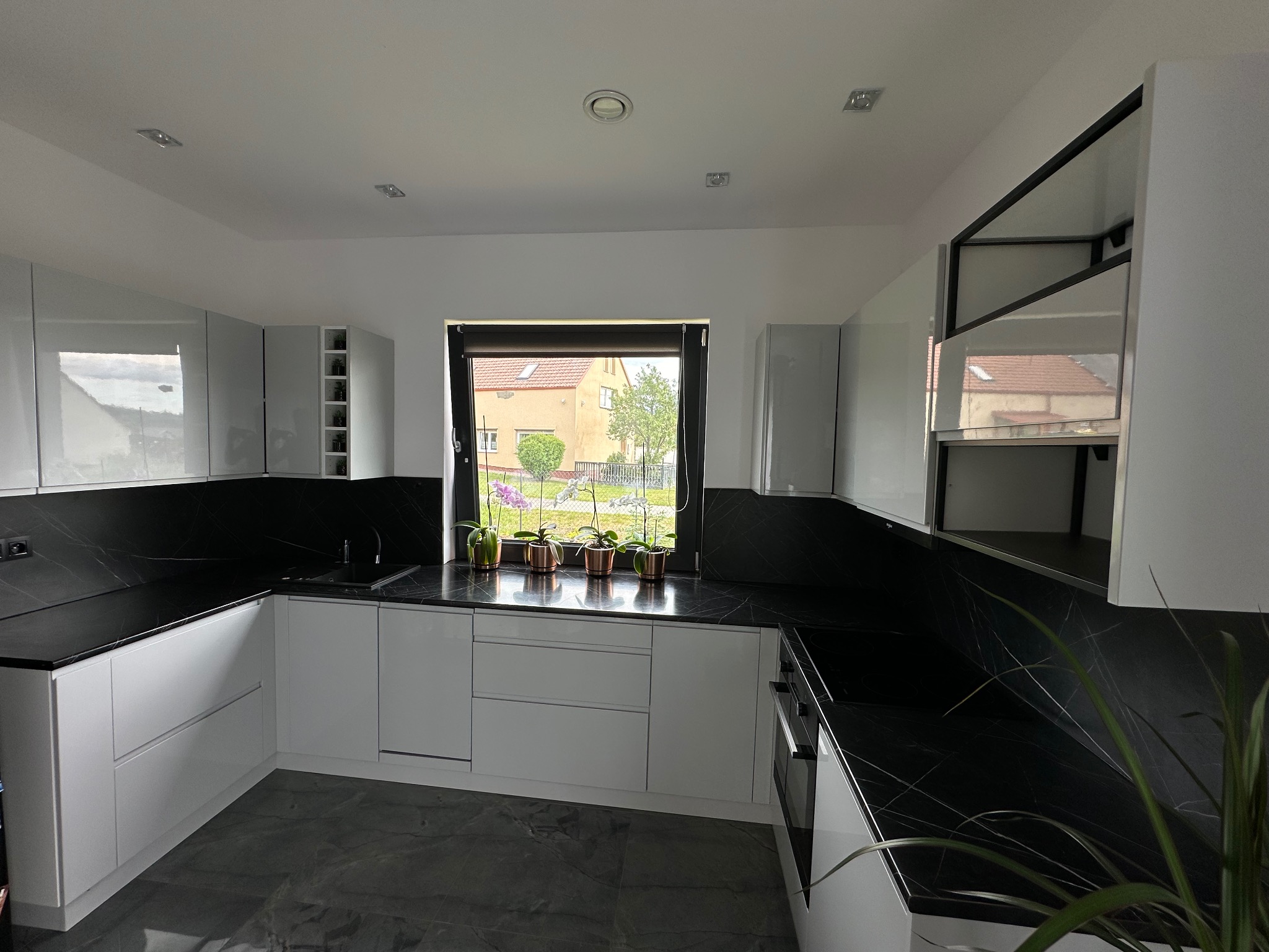 Nowoczesna kuchnia z białymi, lakierowanymi frontami i czarnym blatem z marmurkowym wzorem, widok na okno z roślinami doniczkowymi i zabudowę kuchenną z otwartymi półkami.