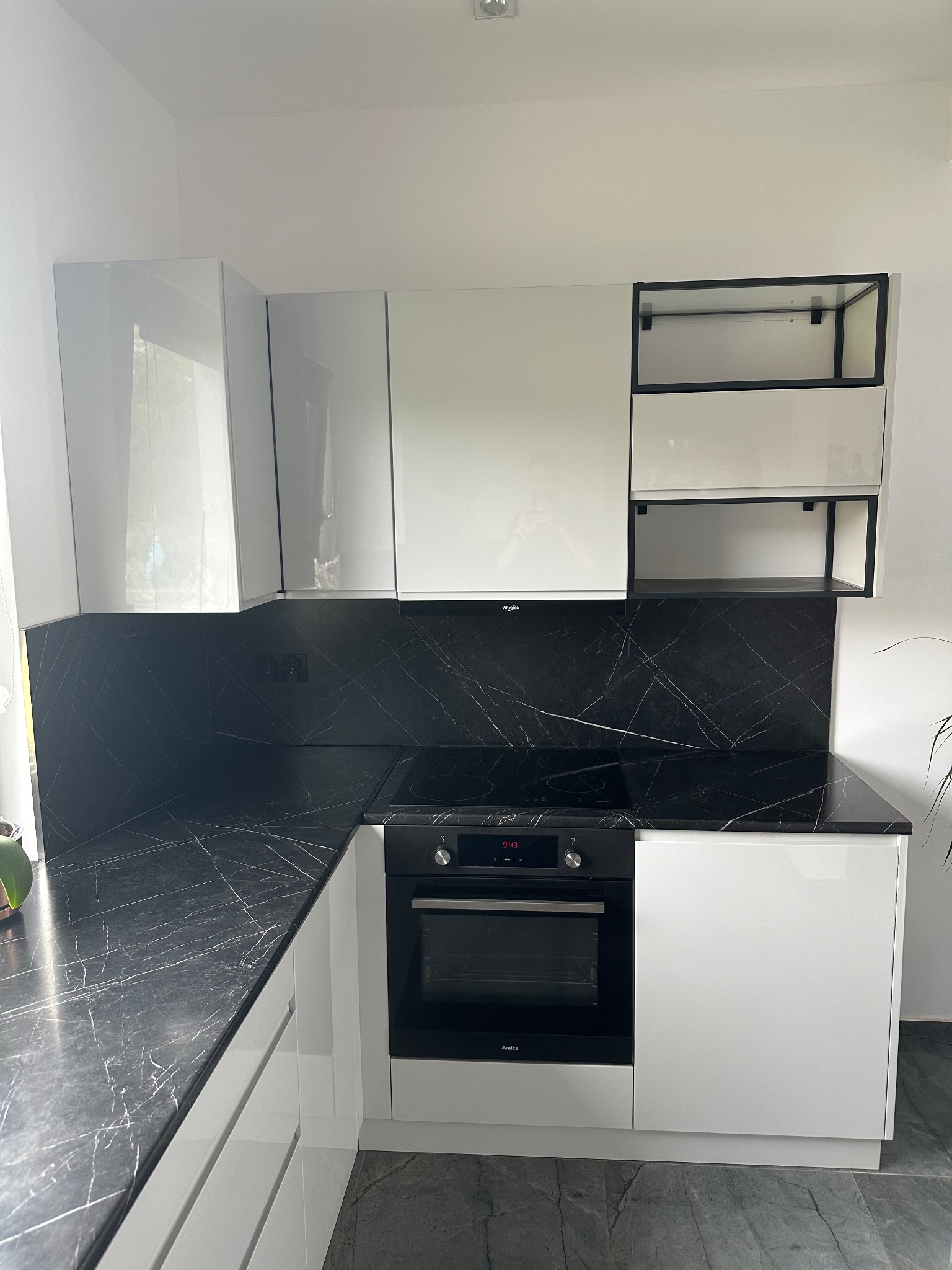 Biała kuchnia z czarnym blatem imitującym marmur, zabudowa górna z szafkami z połyskiem i otwartymi półkami z czarną ramą, piekarnik Amica w zabudowie.