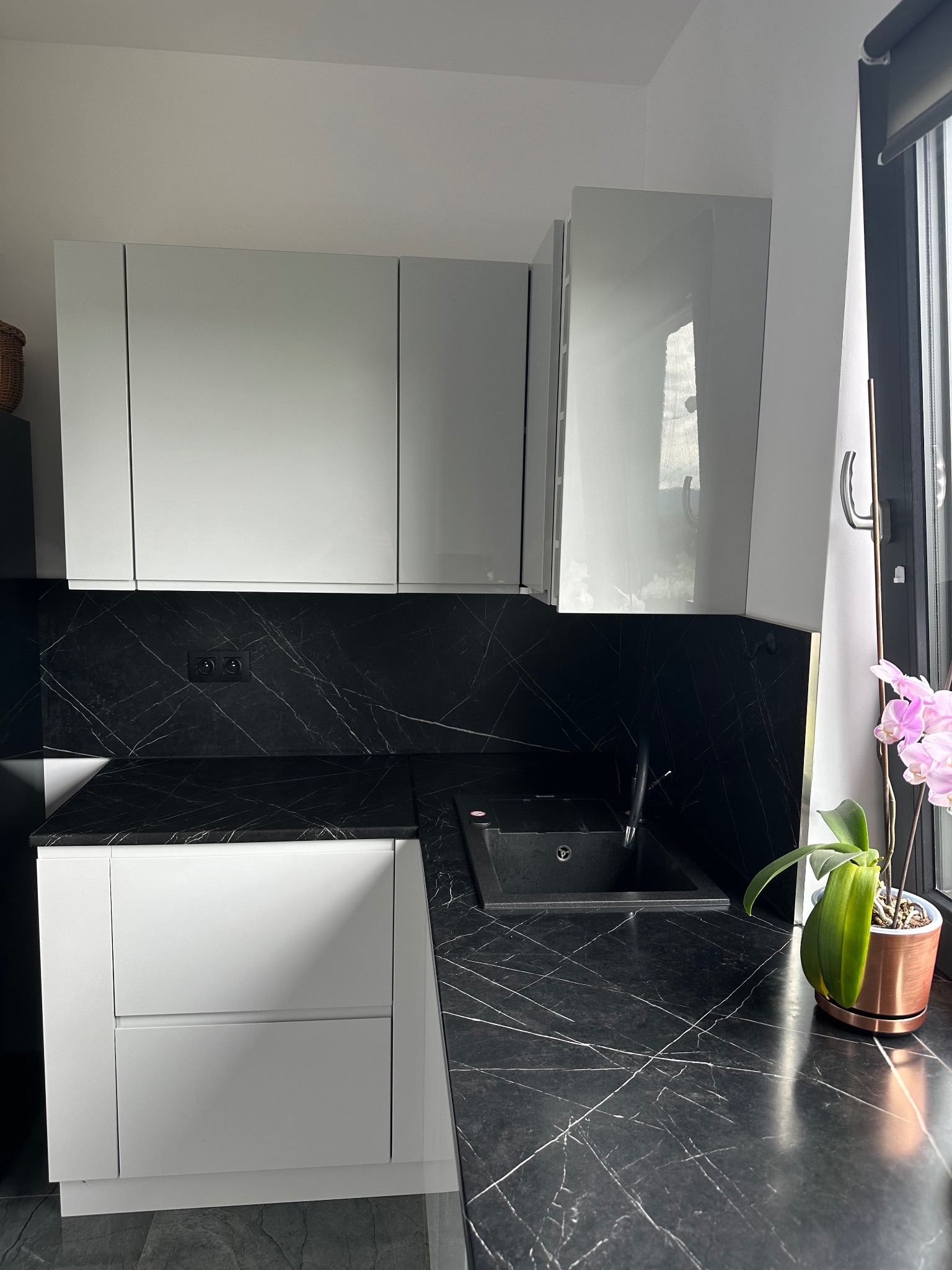 Nowoczesna kuchnia z białymi szafkami i czarnym blatem imitującym marmur, zlew z czarnego granitu, storczyk w miedzianej doniczce przy oknie.