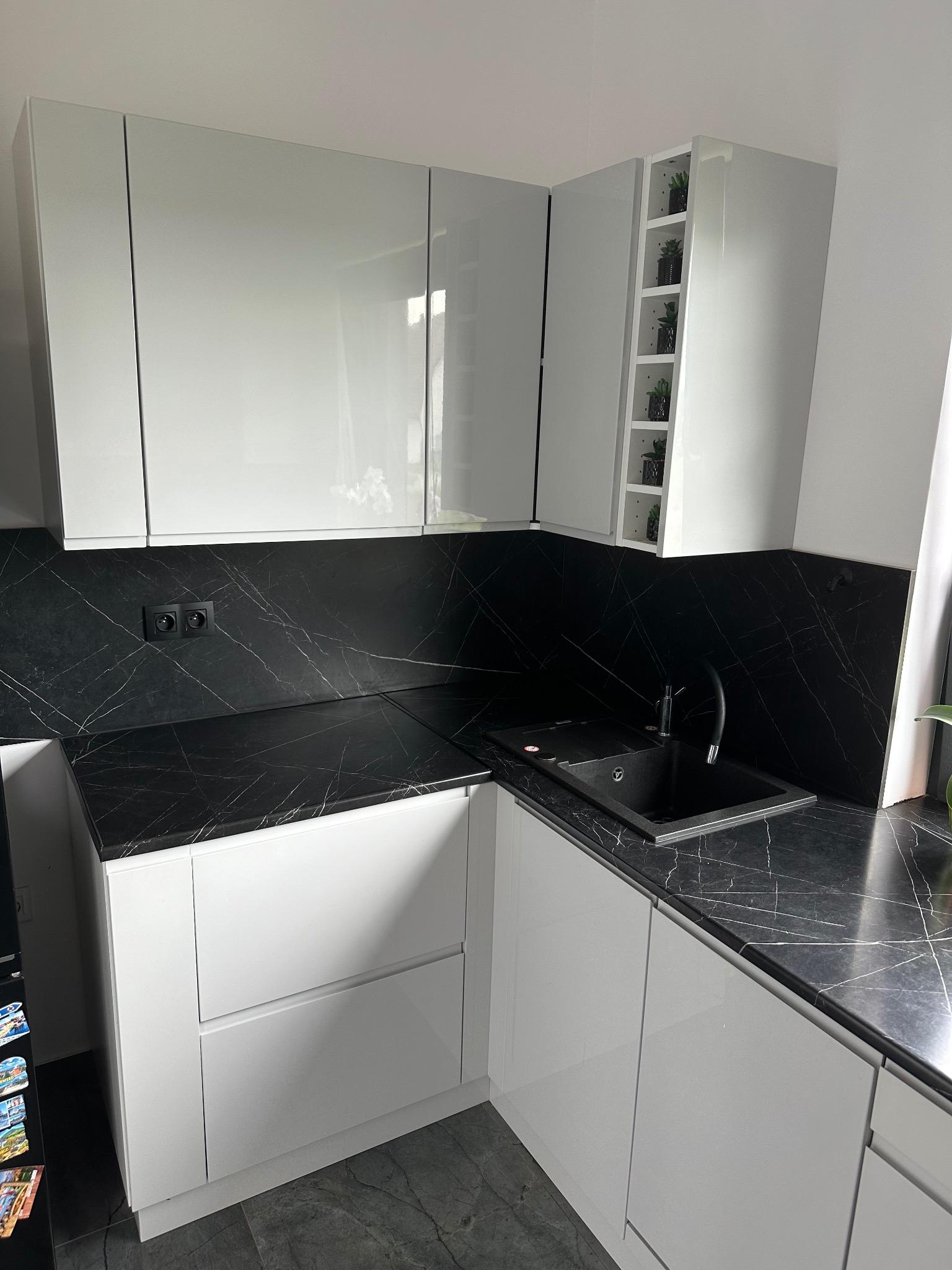 Biała, nowoczesna kuchnia z czarnym blatem o wzorze marmuru i czarnym zlewem, uzupełniona o półki z roślinami.