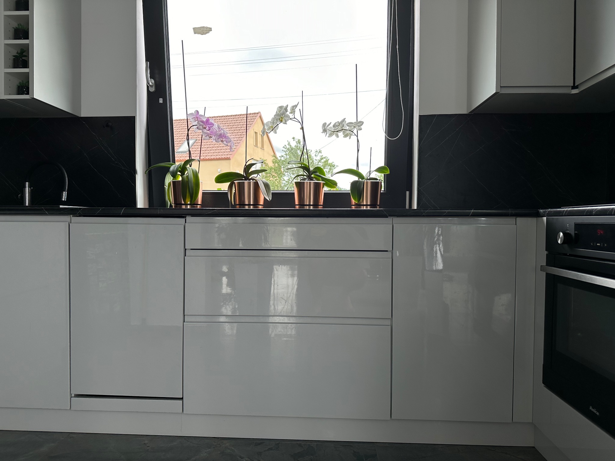 Biała, lakierowana kuchnia z czarnym blatem i backsplashiem imitującym marmur, z rzędem orchidei w miedzianych doniczkach na tle okna.