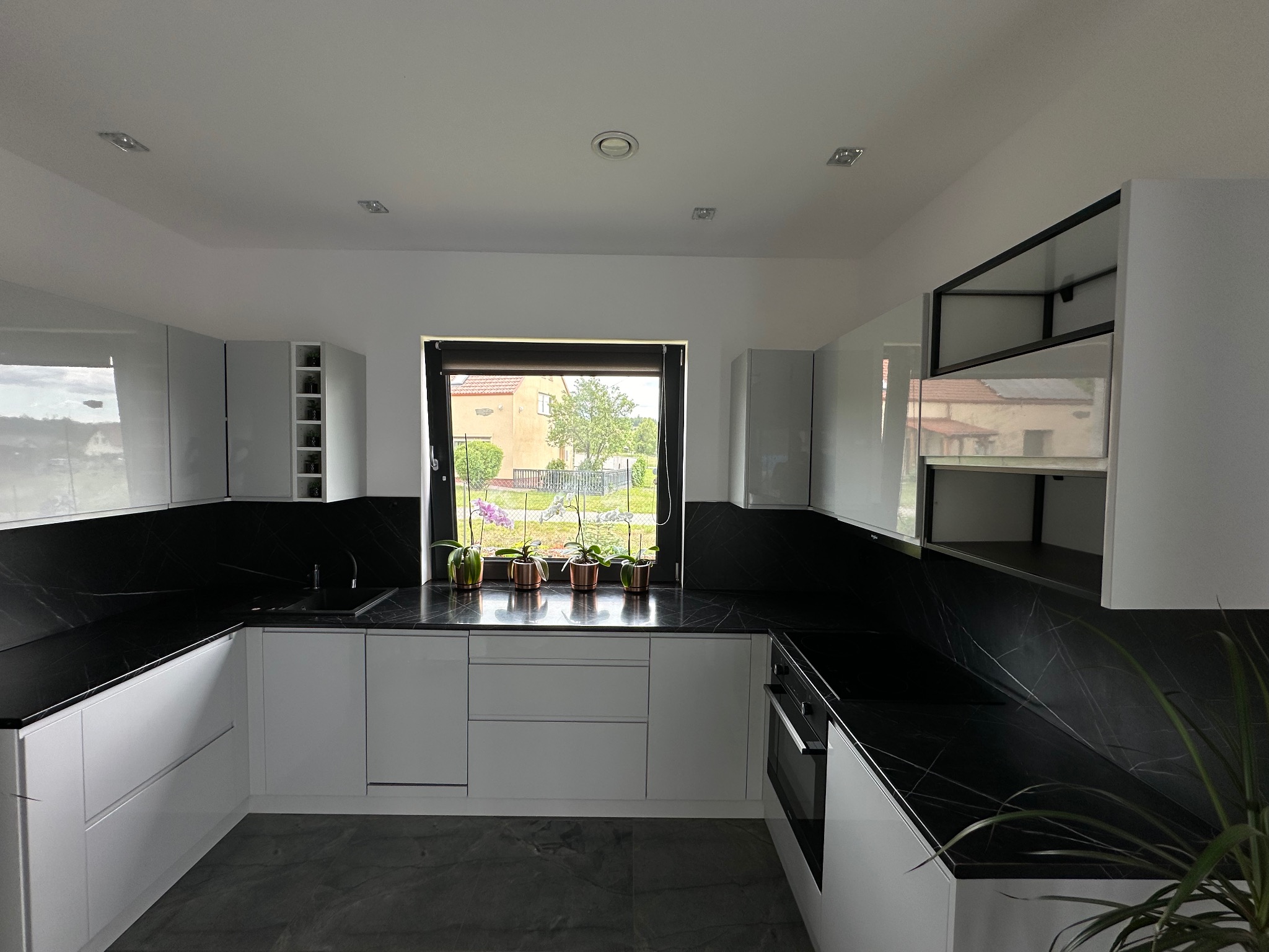 Nowoczesna, jasna kuchnia z białymi, lakierowanymi frontami, czarnym blatem imitującym marmur, szarą podłogą i oknem z widokiem na dom i ogród.