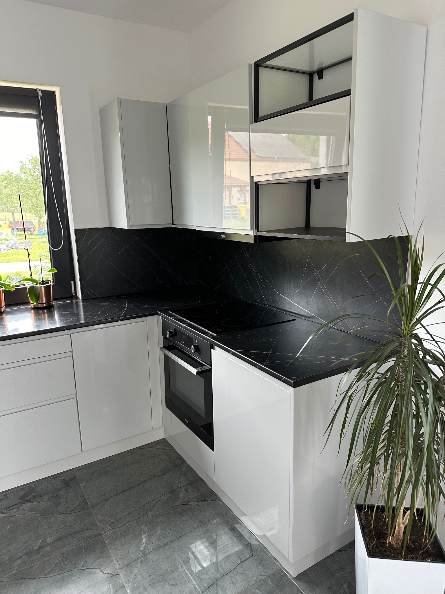 Biała kuchnia z czarnym blatem i płytą indukcyjną, uzupełniona rośliną doniczkową w kwadratowej, białej donicy, widok na zewnątrz przez okno.