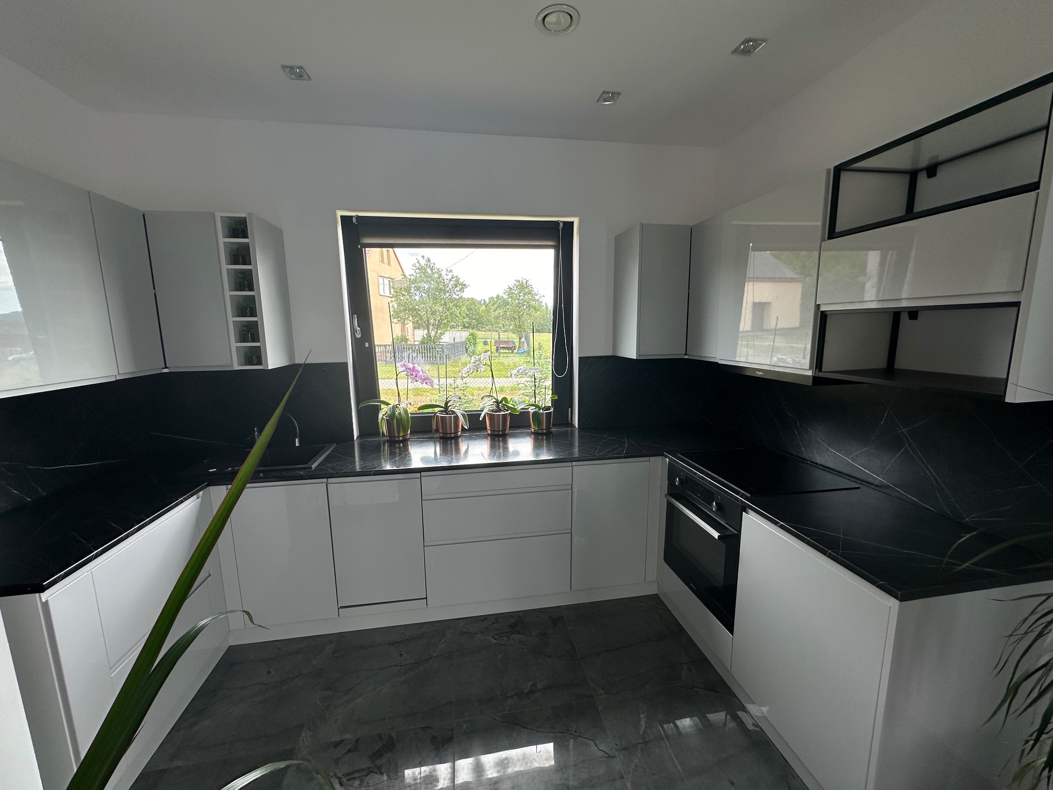Nowoczesna, minimalistyczna kuchnia z białymi, lakierowanymi frontami, czarnym blatem i podłogą imitującą marmur, z widokiem na ogród przez okno, ozdobiona kwiatami w doniczkach.