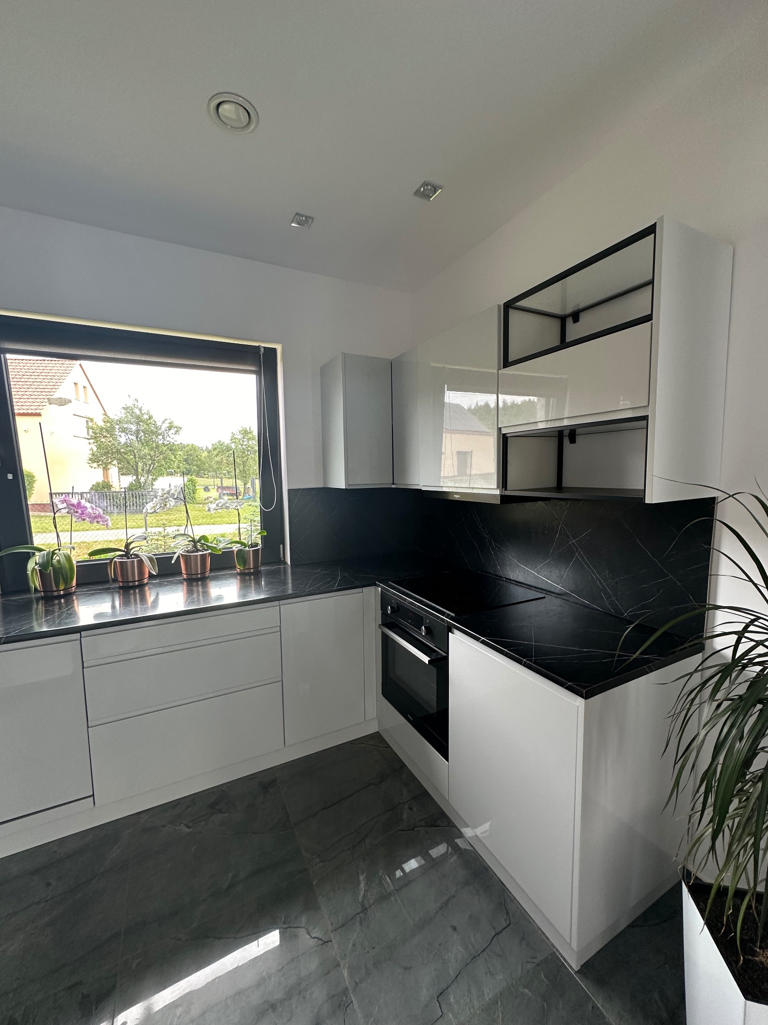 Nowoczesna kuchnia na wymiar z białymi, lakierowanymi frontami, czarnym blatem z marmurowym wzorem i otwartymi półkami z czarnymi ramami, widok na ogród przez okno z kwiatami.