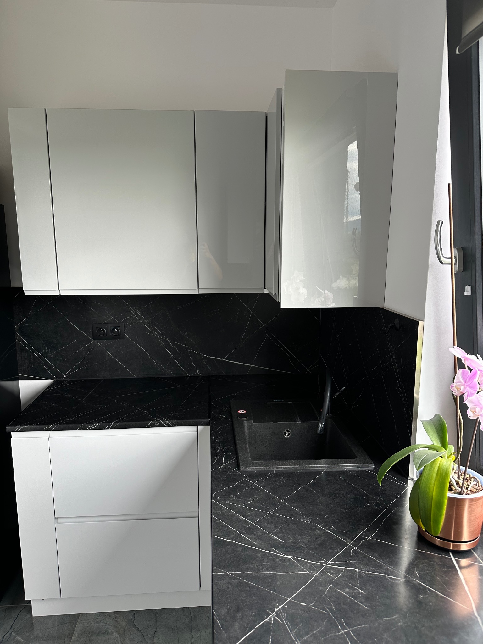 Nowoczesna kuchnia z białymi szafkami i blatem z czarnego marmuru, czarny zlew, kwitnąca orchidea w miedzianej doniczce przy oknie.