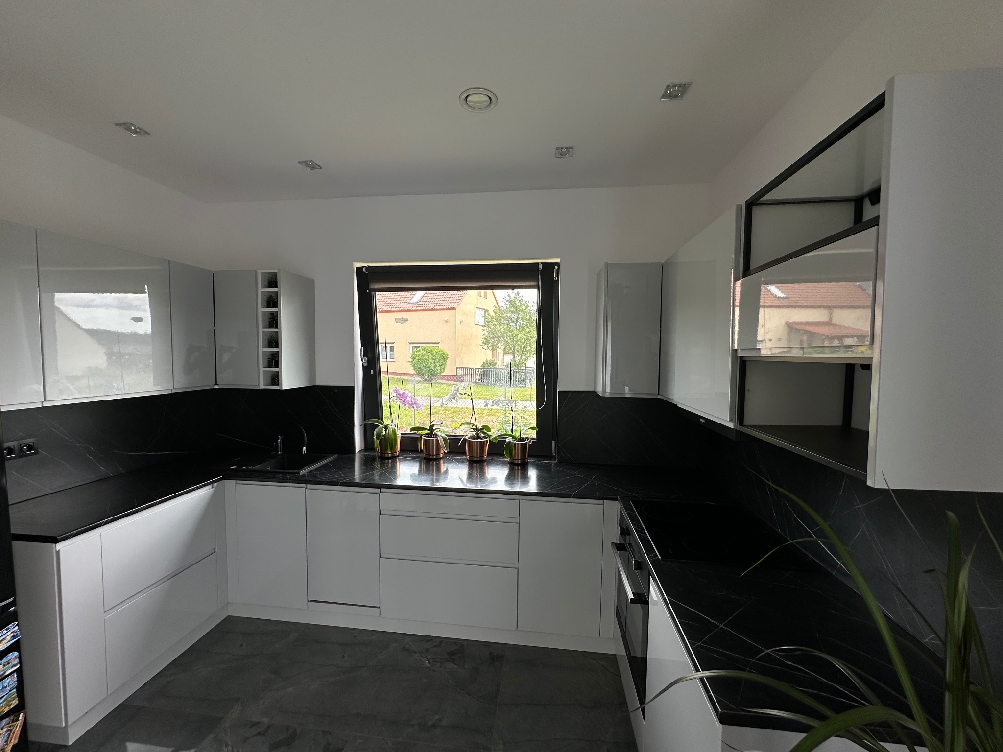 Nowoczesna kuchnia z białymi, lakierowanymi frontami i czarnym blatem imitującym marmur, z oknem w tle i doniczkami z kwiatami na parapecie.