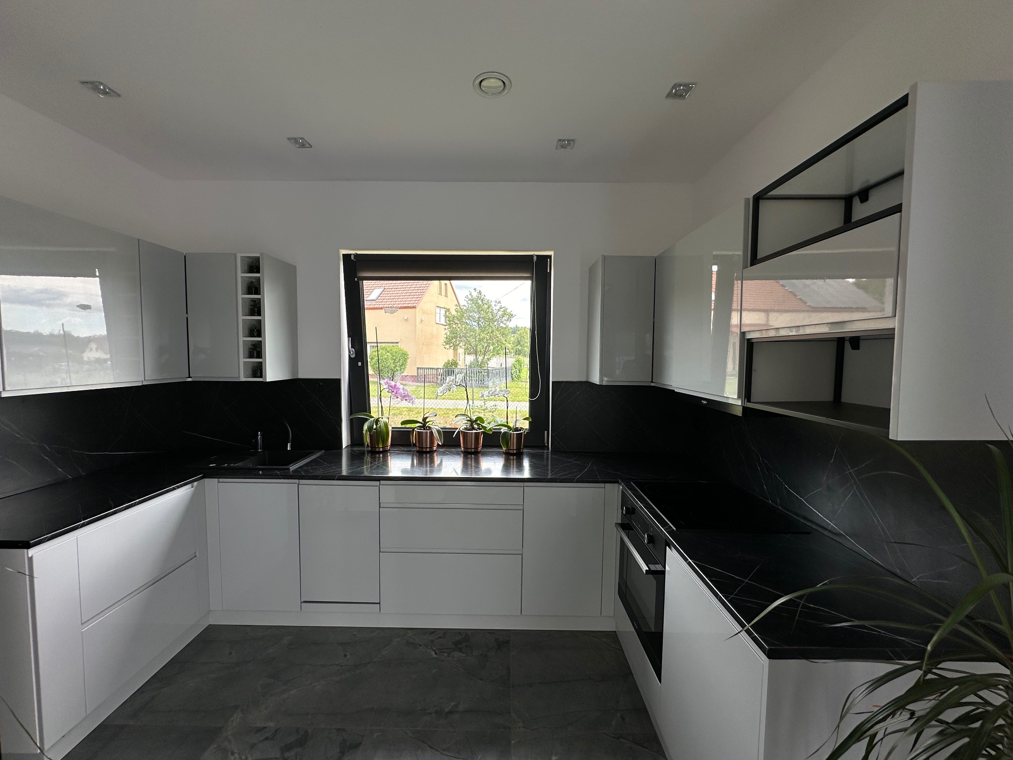 Nowoczesna kuchnia z białymi, lakierowanymi frontami, czarnym blatem z marmurowym wzorem i widokiem na ogród przez okno, gdzie stoją doniczki z kwiatami.