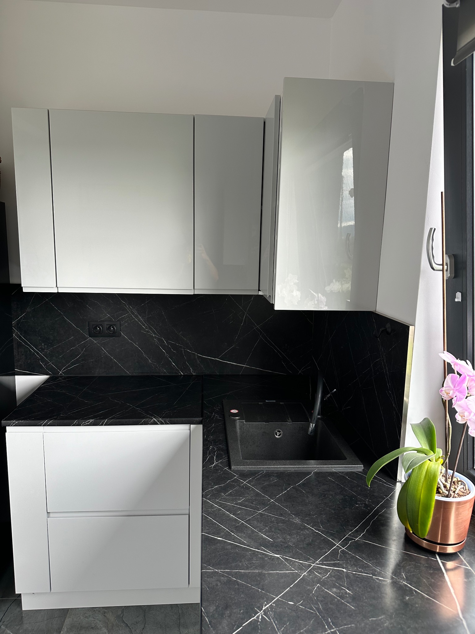 Nowoczesna kuchnia z białymi, błyszczącymi szafkami, blatem i ścianą w czarnym marmurze, czarny zlew i różowy storczyk w miedzianej doniczce przy oknie.