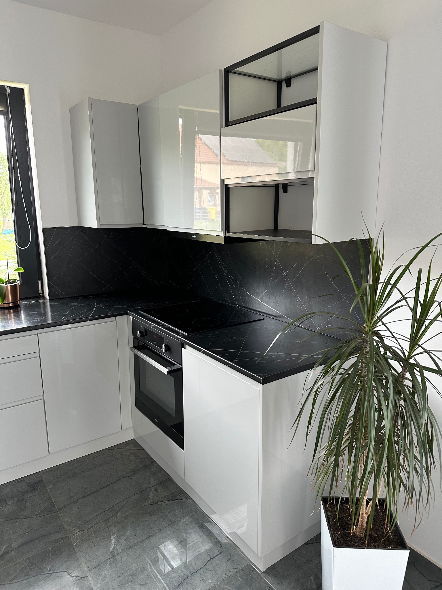 Biała, nowoczesna kuchnia z czarnym blatem imitującym marmur, płyta indukcyjna i piekarnik w zabudowie, szare płytki podłogowe, roślina doniczkowa w białej donicy.