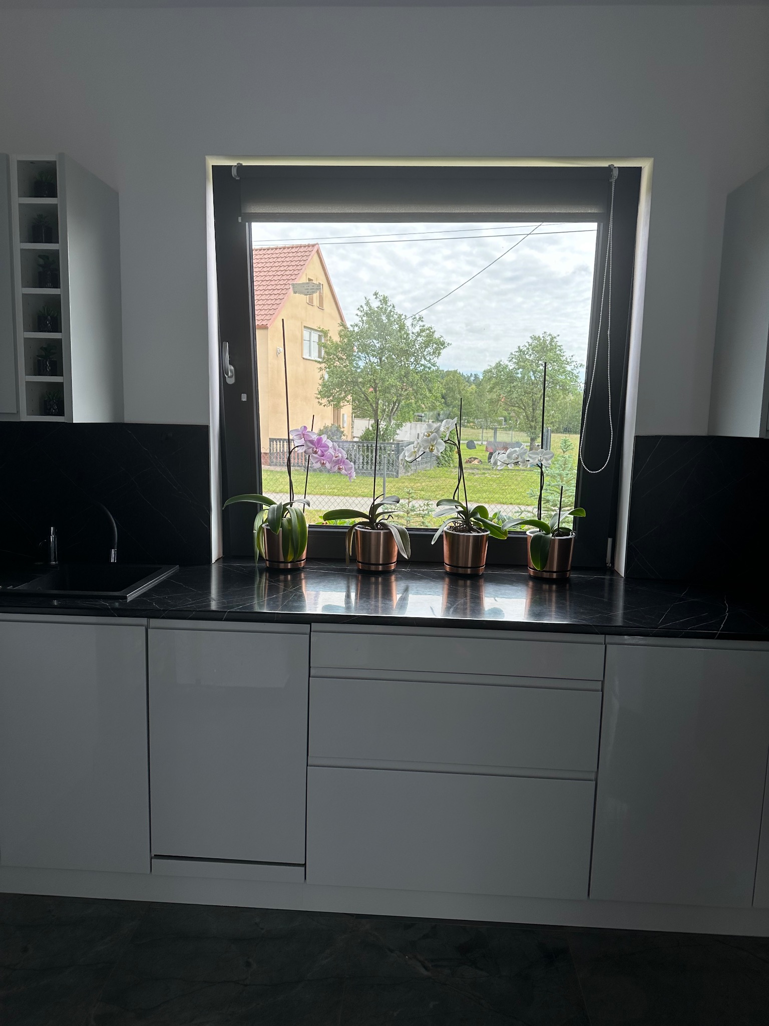 Biała kuchnia z czarnym blatem, cztery orchidee w miedzianych doniczkach na tle okna z widokiem na dom i ogród.