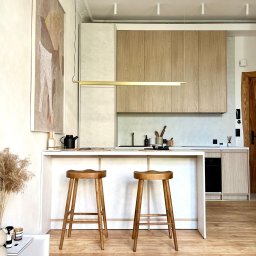 34TH SPÓŁKA Z OGRANICZONĄ ODPOWIEDZIALNOŚCIĄ - Jasna kuchnia z drewnianymi szafkami, białym blatem wyspy i dwoma drewnianymi stołkami barowymi; na ścianie abstrakcyjny obraz, a na blacie wyspy czajnik i dekoracje.