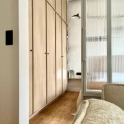 Jasna szafa wnękowa z drewnianymi frontami i złotymi uchwytami, widok z perspektywy wnętrza, z fragmentem fotela na pierwszym planie i ryflowaną szklaną ścianą w tle.