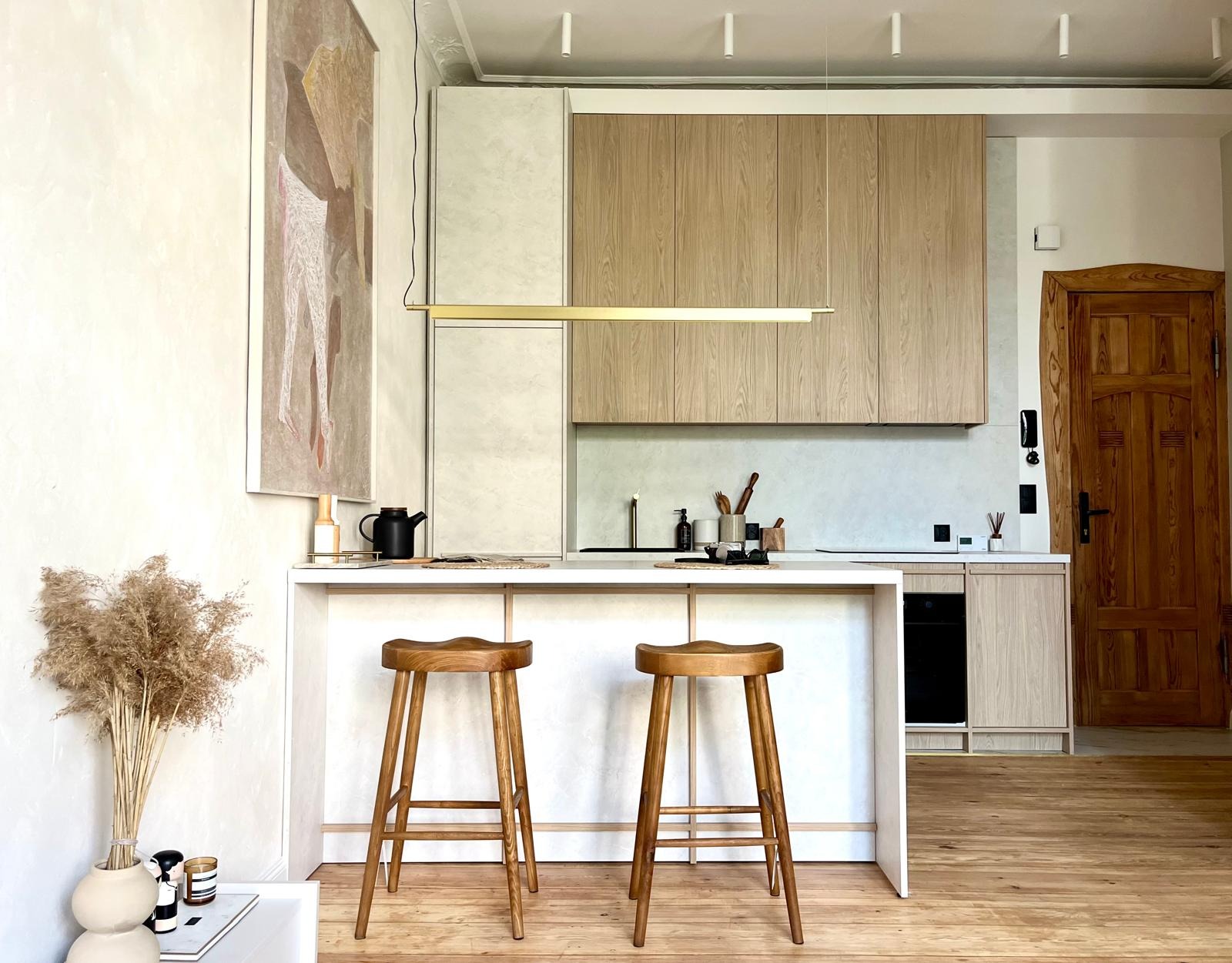 Jasna kuchnia z drewnianymi szafkami, białym blatem wyspy i dwoma drewnianymi stołkami barowymi; na ścianie abstrakcyjny obraz, a na blacie wyspy czajnik i dekoracje.