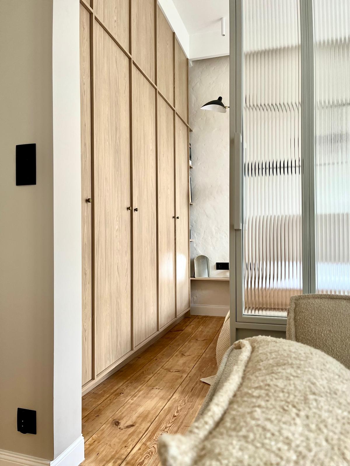 Jasna szafa wnękowa z drewnianymi frontami i złotymi uchwytami, widok z perspektywy wnętrza, z fragmentem fotela na pierwszym planie i ryflowaną szklaną ścianą w tle.