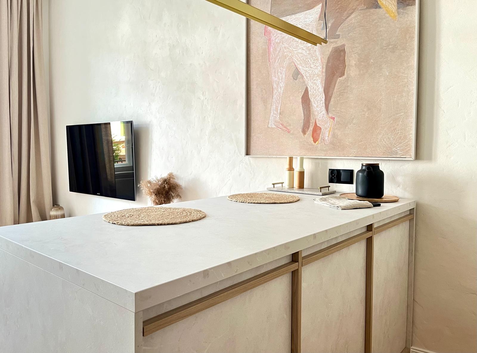 Minimalistyczna wyspa kuchenna z białym blatem i beżowymi frontami, okrągłe podkładki z trawy morskiej, obraz w jasnych barwach na ścianie, telewizor i dekoracje.