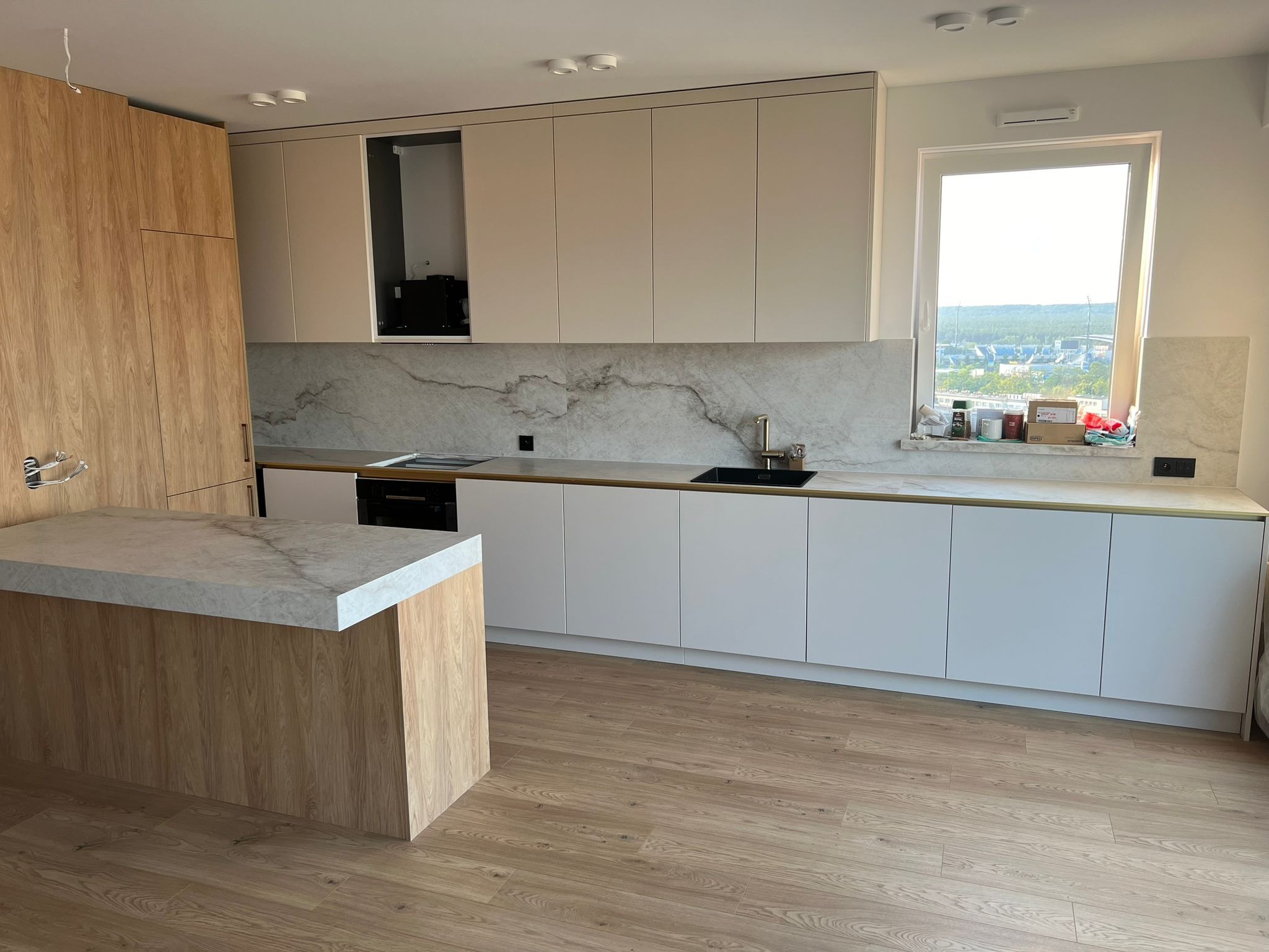 Nowoczesna kuchnia z wyspą, fronty szafek w kolorze złamanej bieli, okładzina ściany nad blatem imitująca marmur, podłoga drewniana.