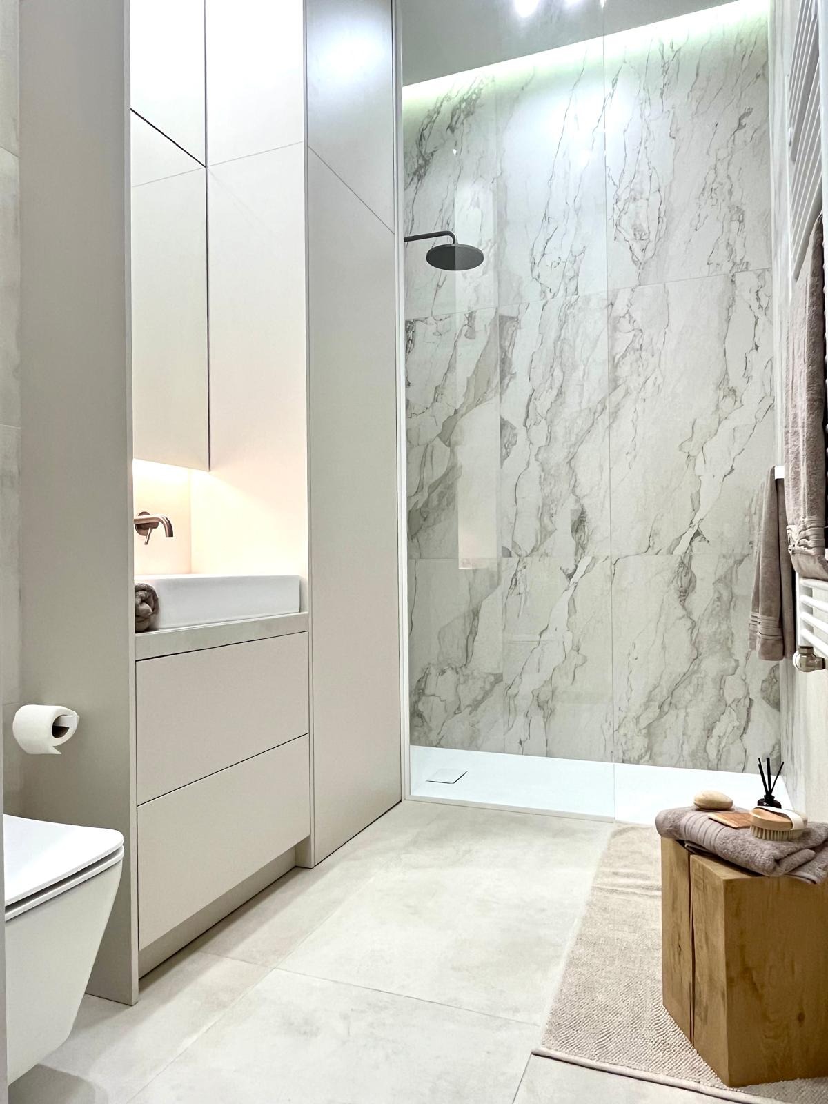 Nowoczesna łazienka w odcieniach szarości z kabiną prysznicową typu walk-in, marmurową ścianą, umywalką w zabudowie i drewnianym stołkiem z ręcznikami.