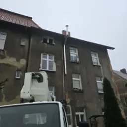 Montaż nowego, metalowego przewodu kominowego na elewacji starego, wielorodzinnego budynku z użyciem podnośnika koszowego. Widoczny fragment kabiny podnośnika oraz elewacja budynku z oknami.