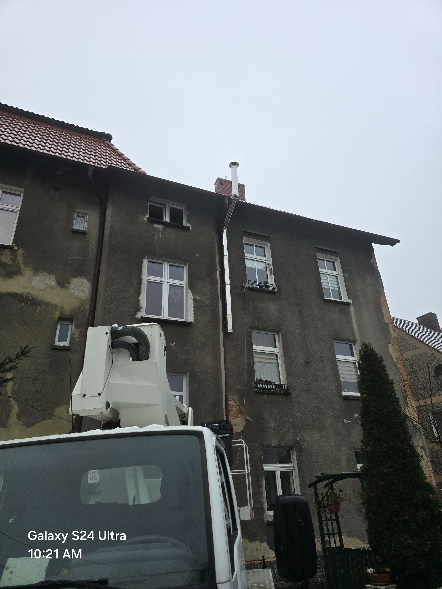 Montaż nowego, metalowego przewodu kominowego na elewacji starego, wielorodzinnego budynku z użyciem podnośnika koszowego. Widoczny fragment kabiny podnośnika oraz elewacja budynku z oknami.