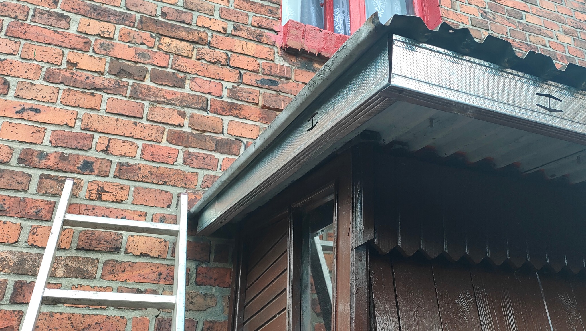 Metalowa obróbka blacharska okapu nad oknem, widoczna drabina oparta o ceglaną ścianę budynku.