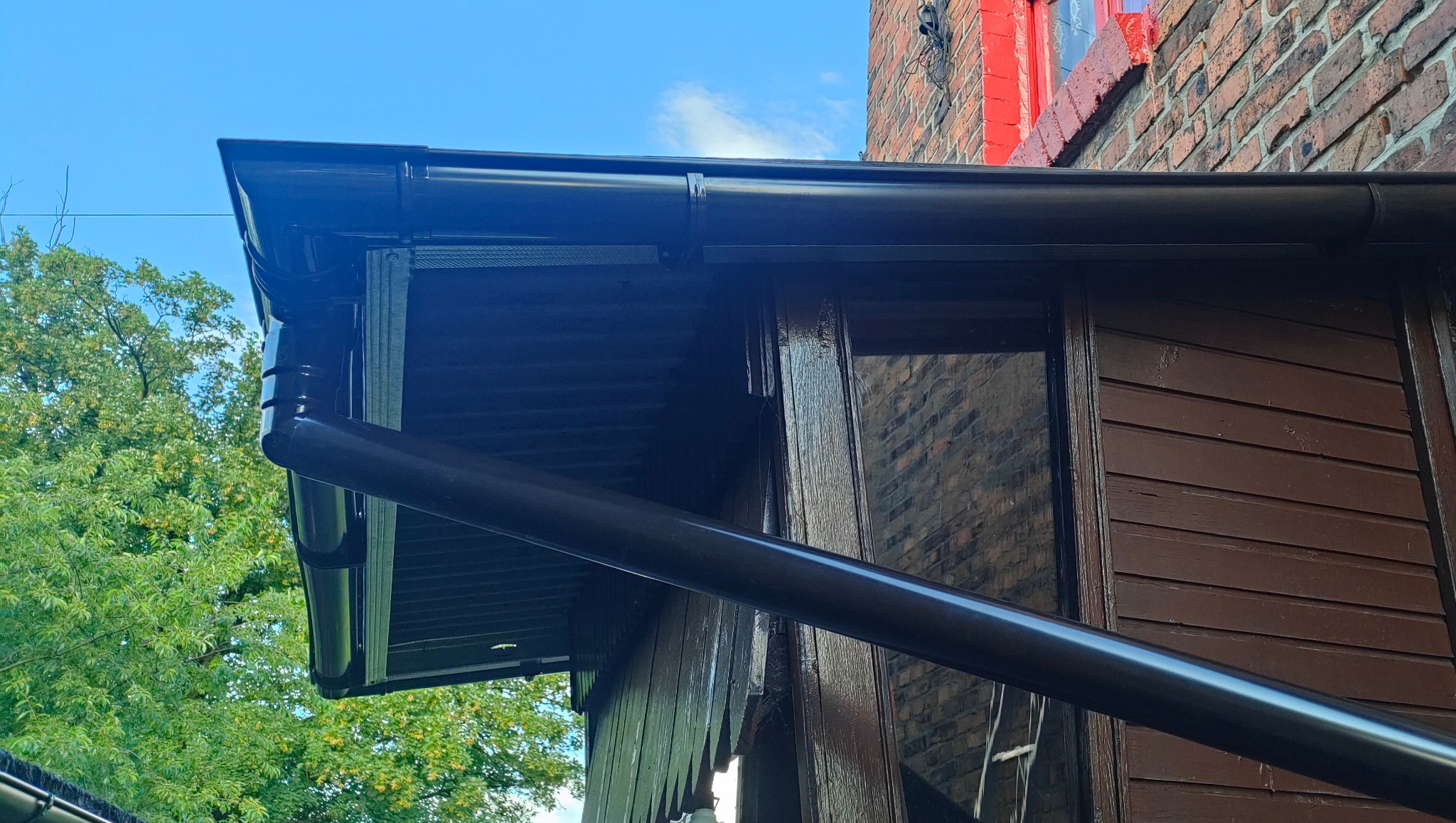 Ciemnobrązowe rynny i rura spustowa zamontowane na drewnianej konstrukcji budynku, z widoczną ceglaną ścianą sąsiedniego budynku w tle.