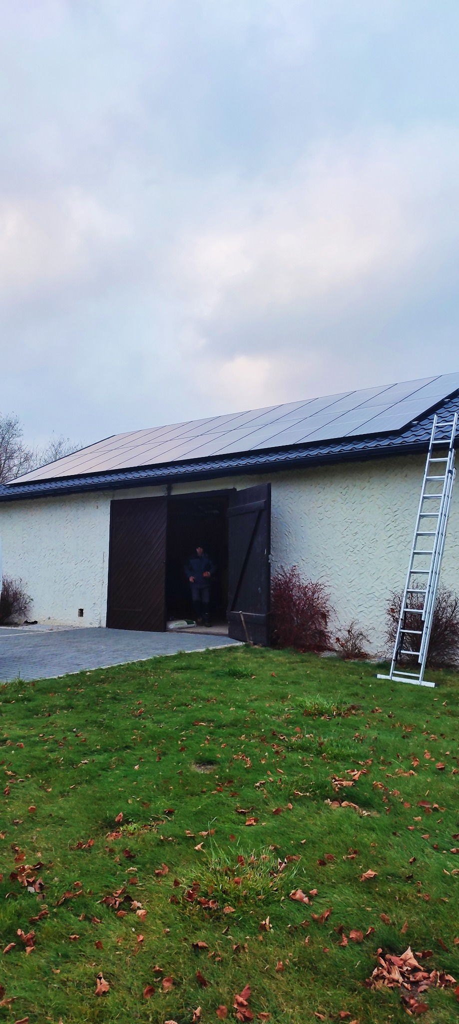 Biały budynek gospodarczy z panelami słonecznymi na dachu, drabina oparta o ścianę, otwarte dwuskrzydłowe drzwi ukazujące sylwetkę osoby w środku.