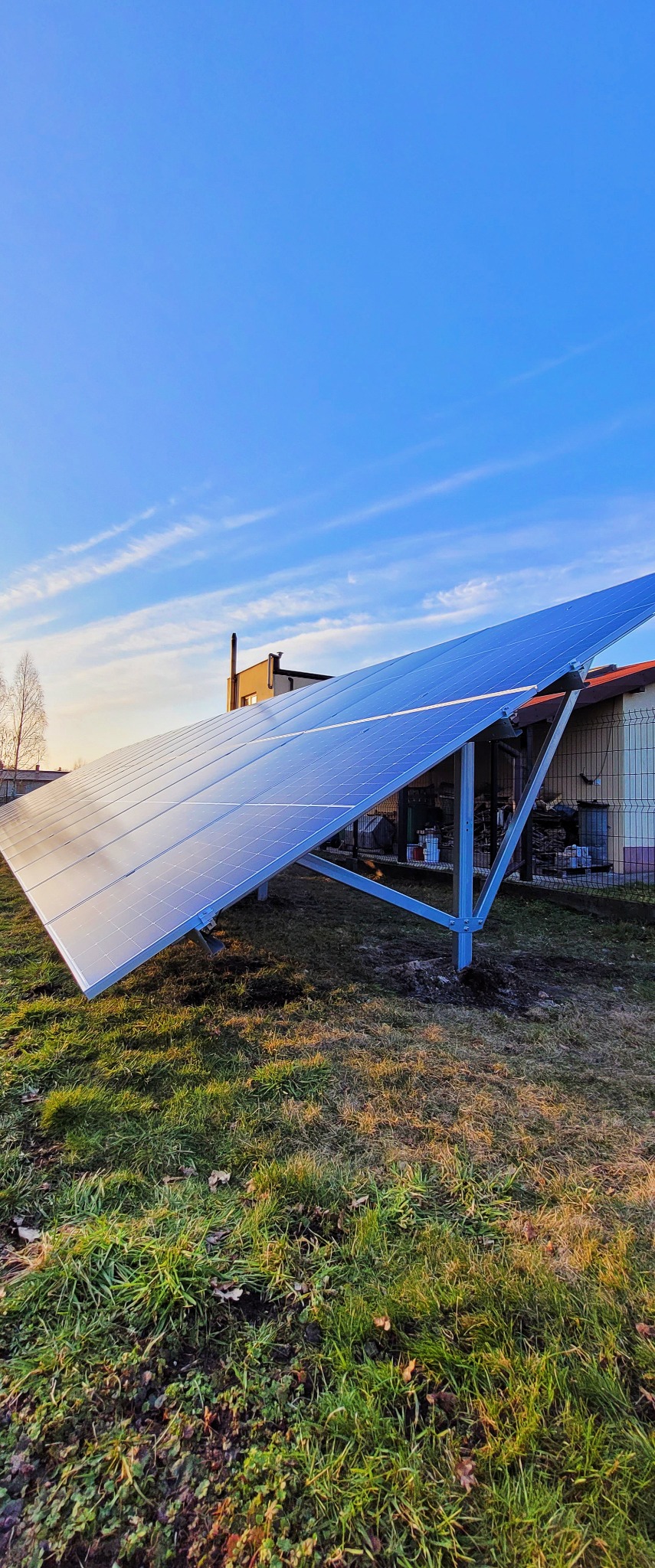 Panel fotowoltaiczny na metalowej konstrukcji, zainstalowany na trawiastym terenie przy domu jednorodzinnym, błękitne niebo z delikatnymi chmurami w tle.