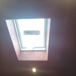 Andrzej Puchała PA&Synowie - Widok z dołu na okno dachowe wbudowane w sufit w kolorze burgundu, z widocznym fragmentem otoczenia na zewnątrz okna.