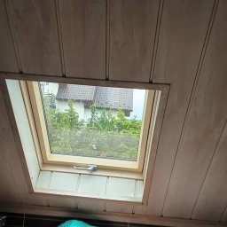 Andrzej Puchała PA&Synowie - Widok z wnętrza poddasza na okno dachowe z widokiem na dom i zieleń, drewniana boazeria na skosach dachu, w dolnej części kadru widoczne przedmioty osobiste.