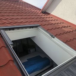 Andrzej Puchała PA&Synowie - Nowo zamontowane okno dachowe z widokiem na wnętrze pokoju zabezpieczonego folią, otoczone czerwoną dachówką i szarymi obróbkami blacharskimi.