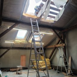 Andrzej Puchała PA&Synowie - Wnętrze poddasza w trakcie wymiany dachu, widoczne drewniane krokwie, izolacja, folia dachowa z widocznymi napisami, dwie drabiny aluminiowe, stos wełny mineralnej, oraz fragment sylwetki...