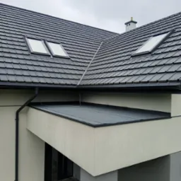 Czarny dach z dachówką i oknami dachowymi, widok z góry. Rynna pionowa po lewej stronie. Fragment elewacji budynku w neutralnym kolorze.