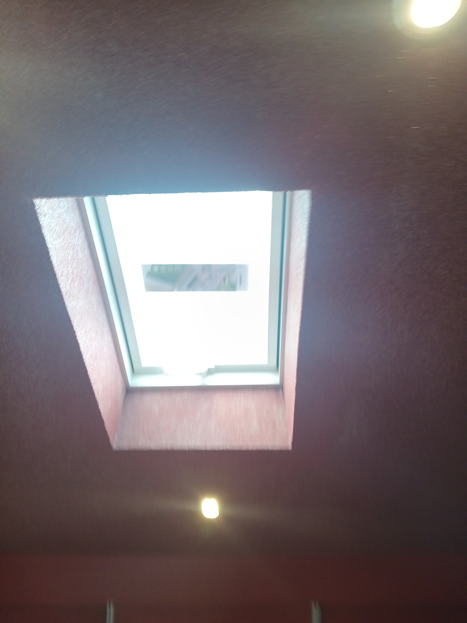 Widok z dołu na okno dachowe wbudowane w sufit w kolorze burgundu, z widocznym fragmentem otoczenia na zewnątrz okna.
