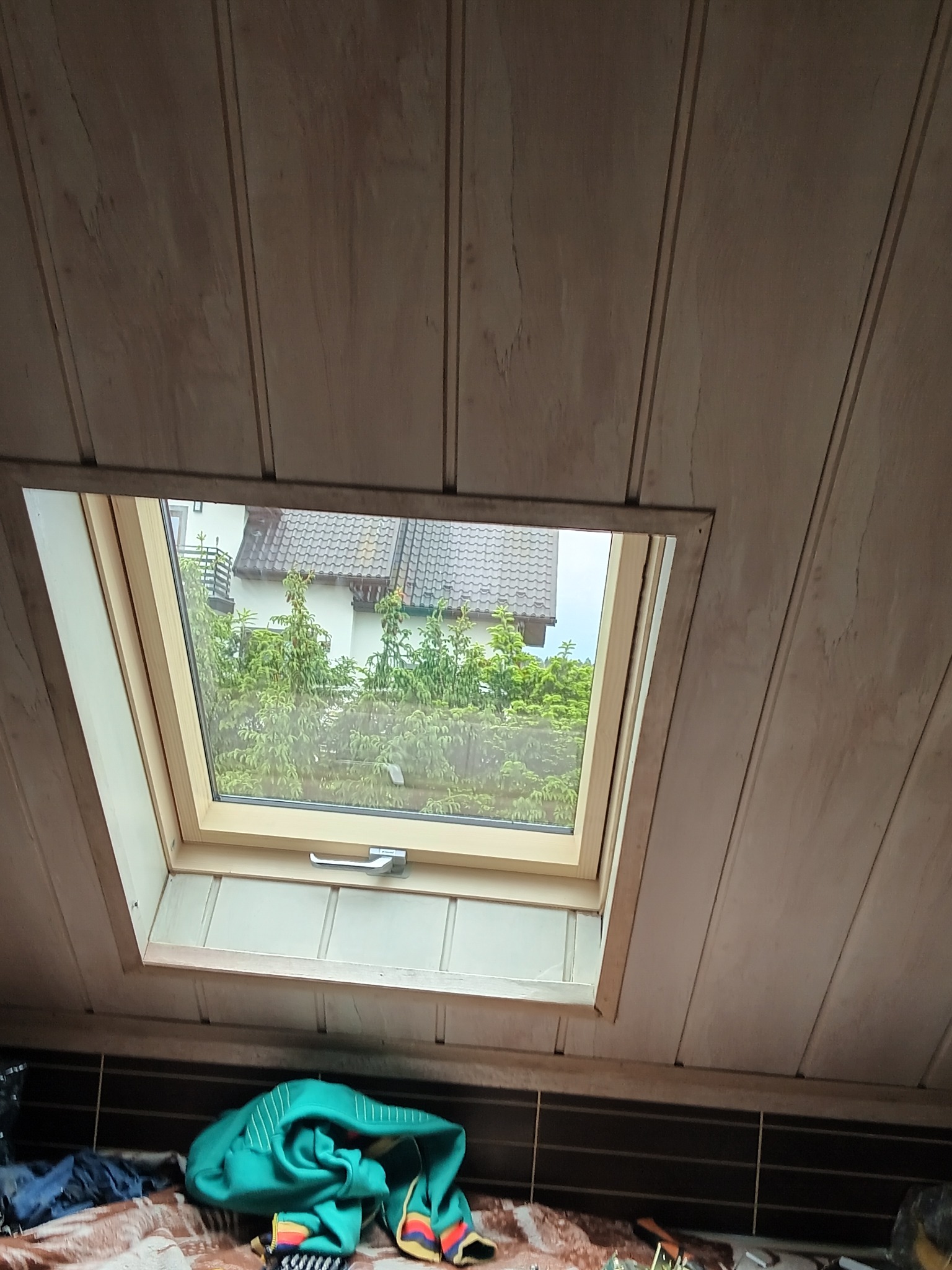 Widok z wnętrza poddasza na okno dachowe z widokiem na dom i zieleń, drewniana boazeria na skosach dachu, w dolnej części kadru widoczne przedmioty osobiste.