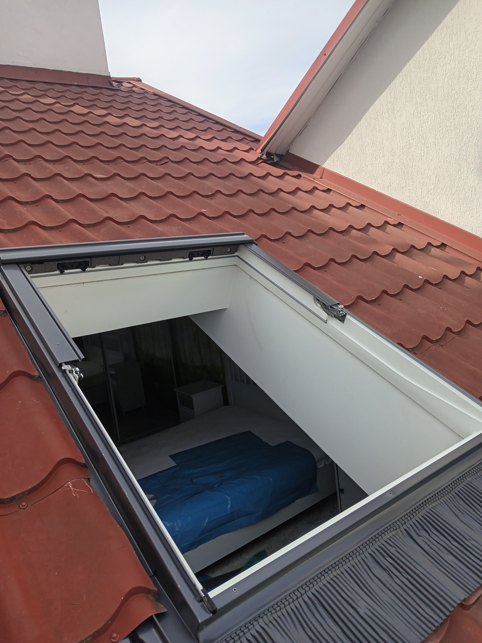 Nowo zamontowane okno dachowe z widokiem na wnętrze pokoju zabezpieczonego folią, otoczone czerwoną dachówką i szarymi obróbkami blacharskimi.