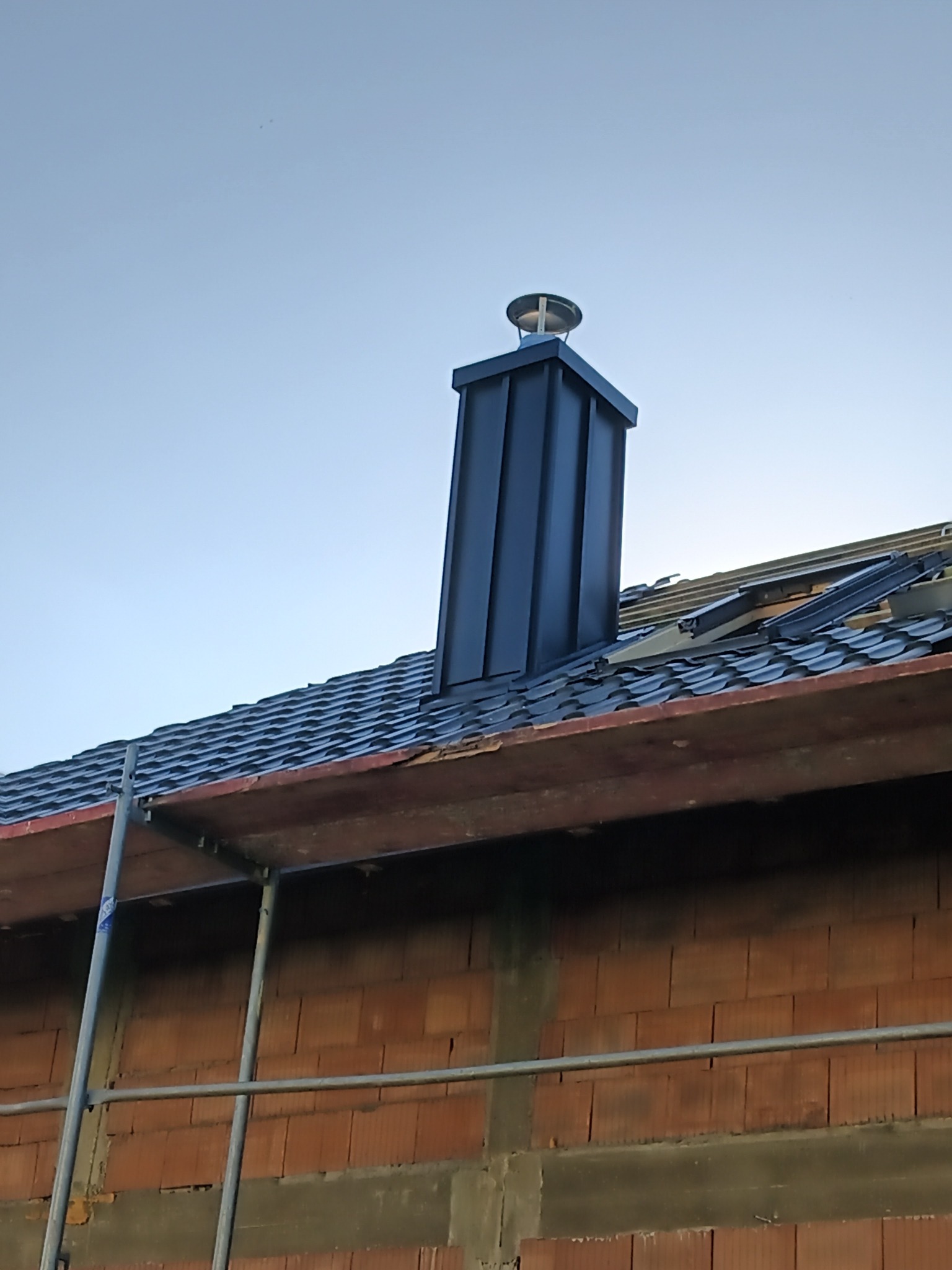 Nowy, czarny komin z metalu na dachu z dachówki ceramicznej, widoczne rusztowanie i fragment muru z cegły.