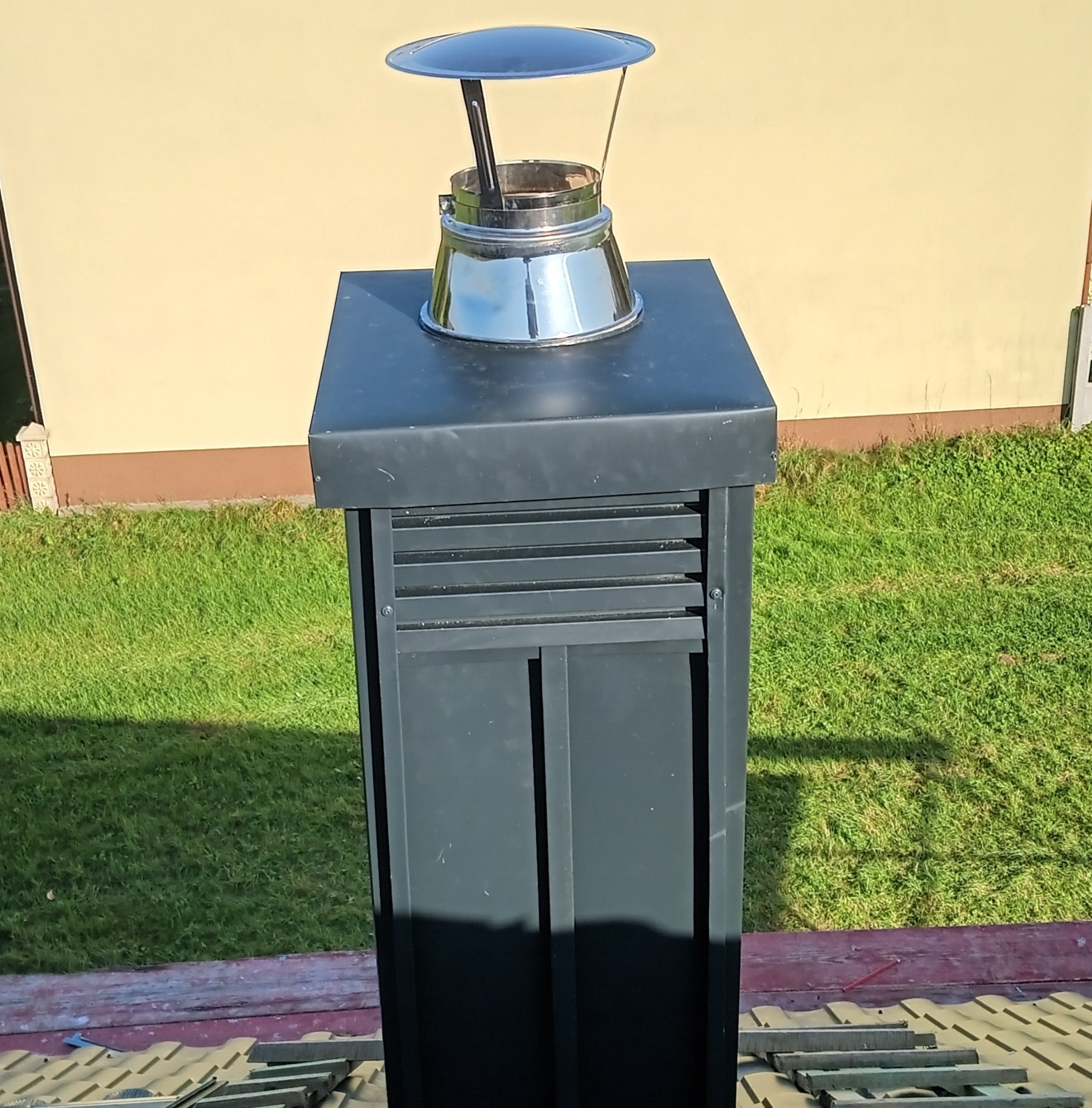 Nowy, czarny komin wentylacyjny z metalowym daszkiem na dachu z dachówki ceramicznej, widok z dołu na tle trawnika i ściany budynku.