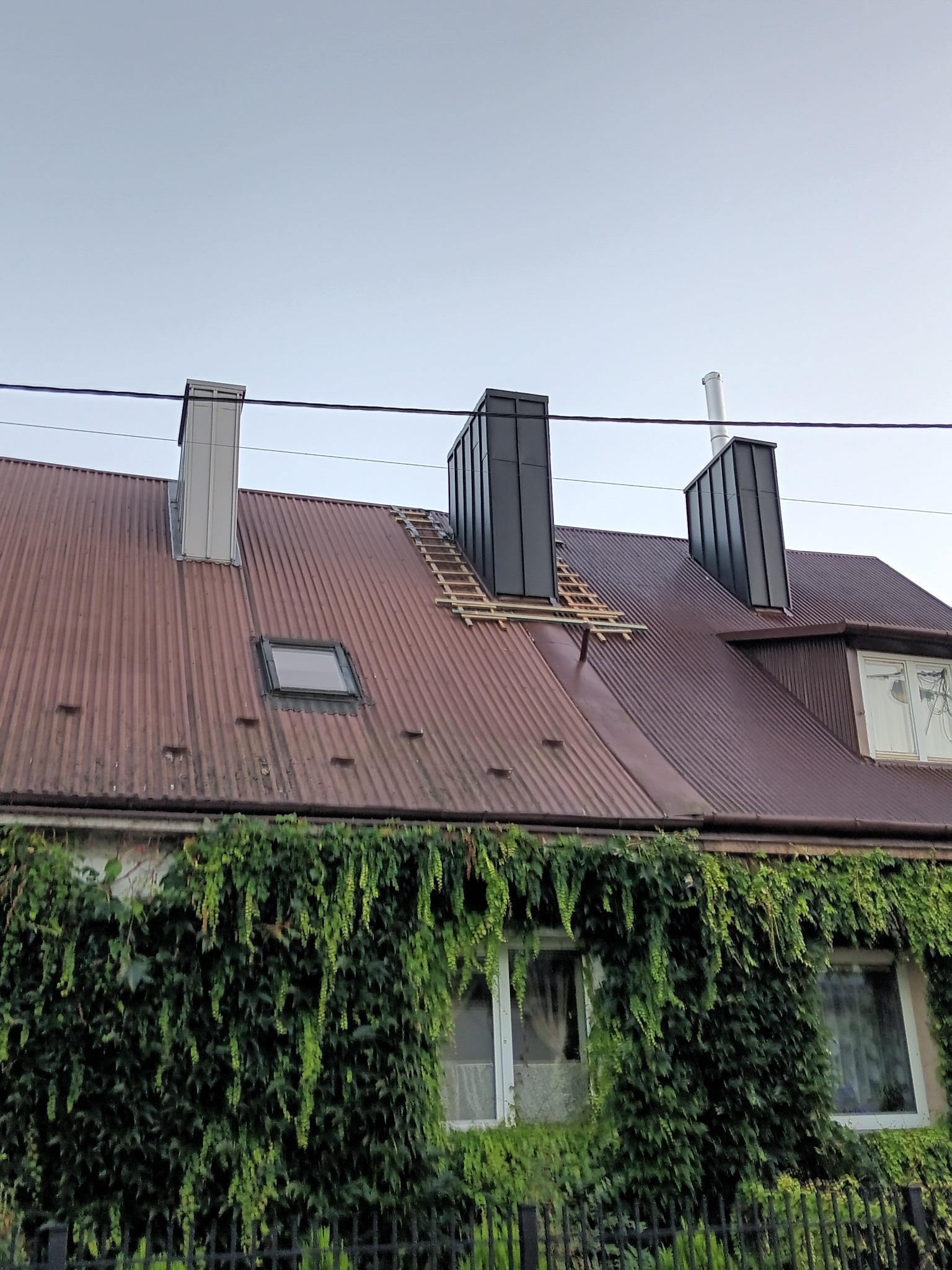 Częściowo wymieniony, pordzewiały dach z blachy falistej z nowymi, czarnymi kominami, widoczne elementy drewniane konstrukcji dachu, okno dachowe i ściana budynku porośnięta bluszczem.