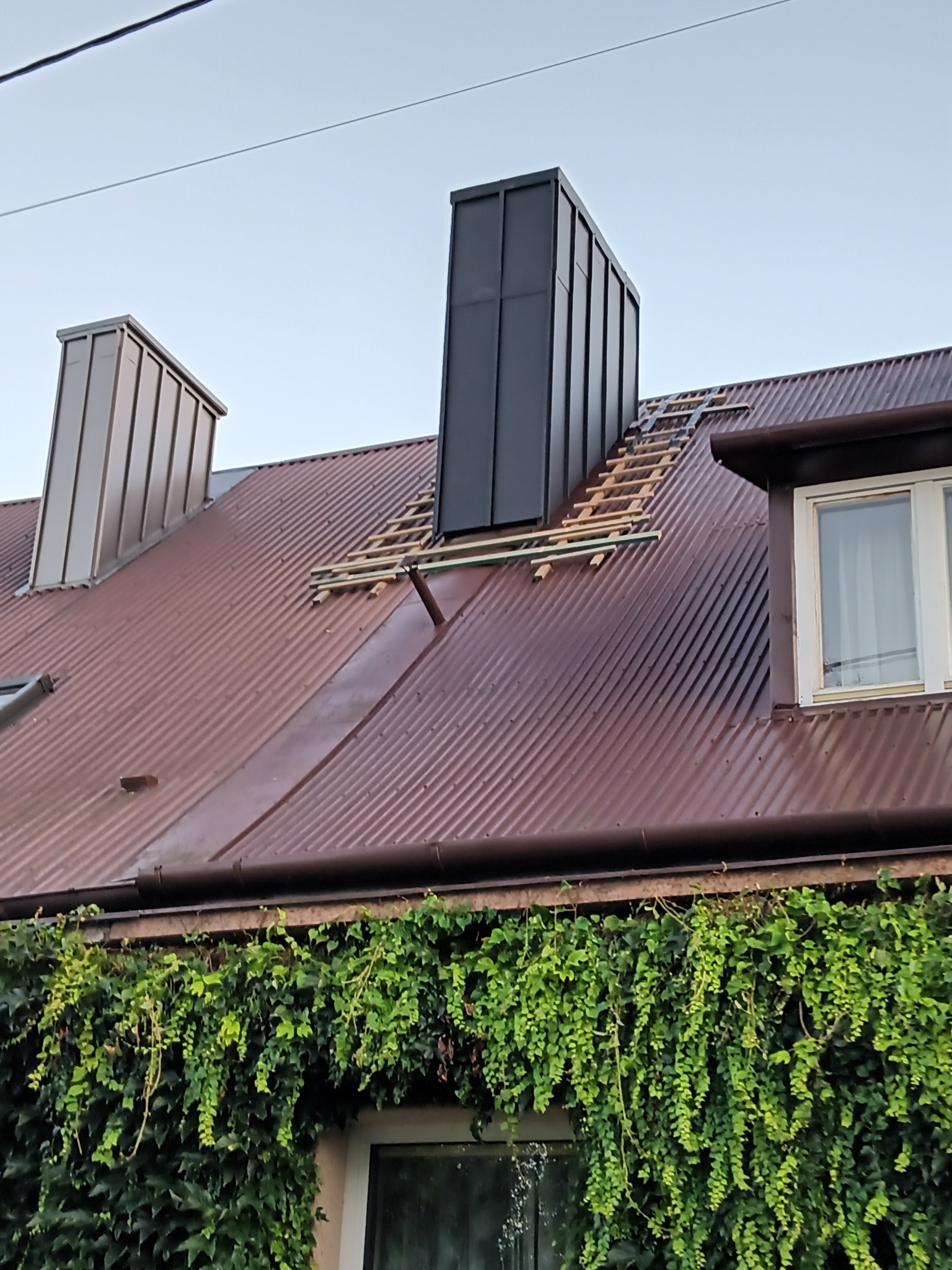 Dom z brązowym dachem z blachy trapezowej, dwa kominy obłożone blachą, jeden w kolorze szarym, drugi w kolorze jasnego brązu, częściowo zdjęte poszycie dachu, widoczne łaty dachowe, elewacja...