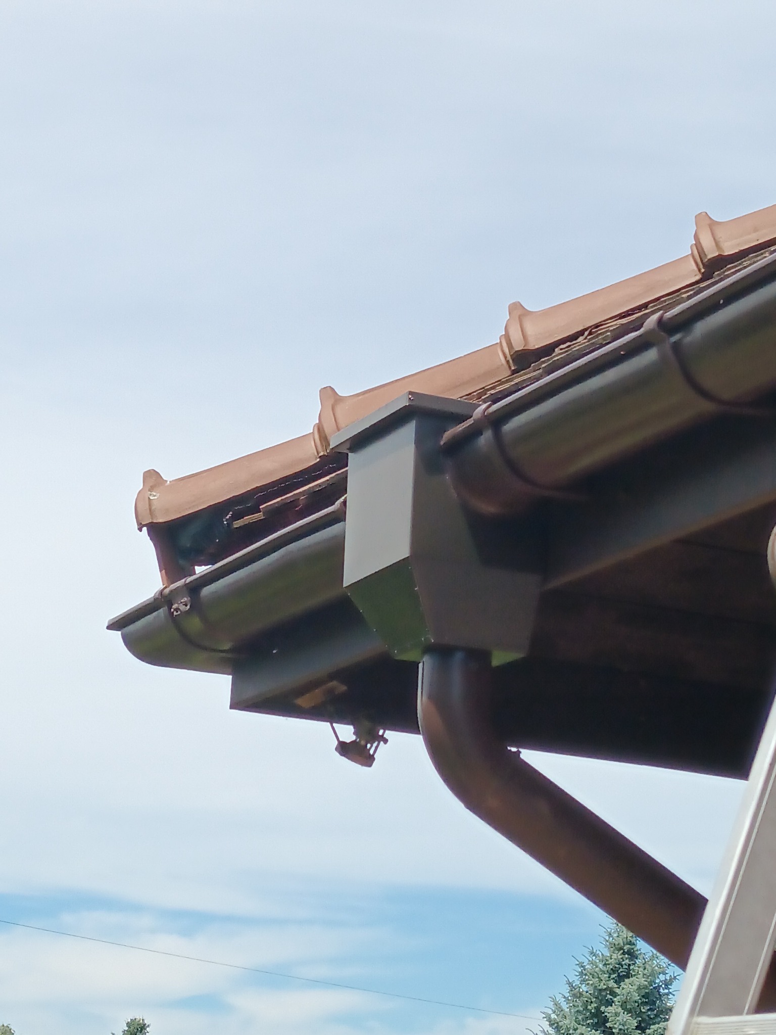 Brązowe rynny dachowe z elementami mocującymi, widoczne z dołu na tle niebieskiego nieba i fragmentu drabiny.