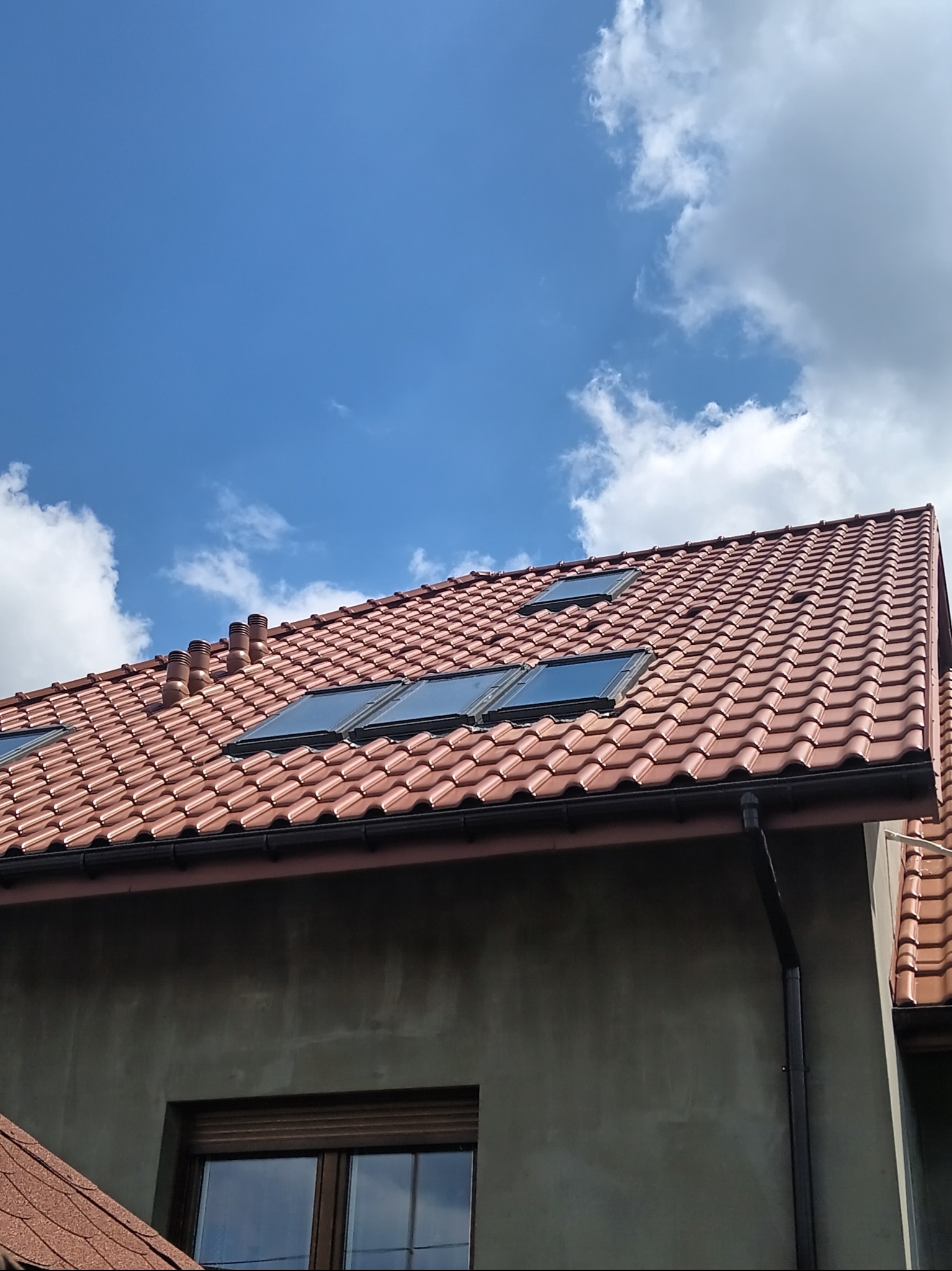 Dom z nowym, czerwonym dachem z dachówki ceramicznej, trzema oknami dachowymi i czterema kominami wentylacyjnymi na tle błękitnego nieba z chmurami.