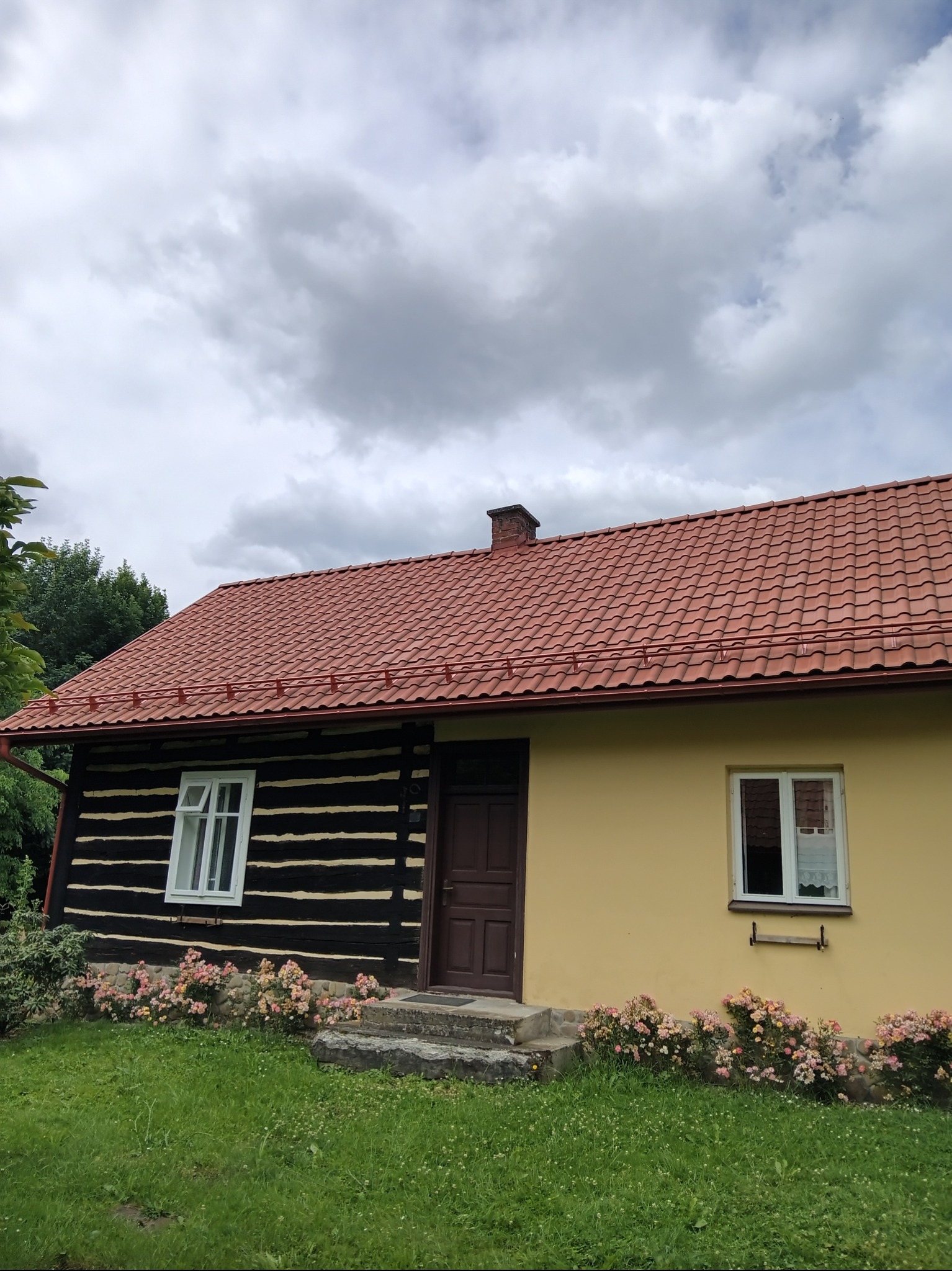 Dom z dwoma różnymi typami elewacji: drewnianą, pomalowaną na czarno z jasnymi belkami, oraz otynkowaną na żółto, z nowym, czerwonym dachem z dachówki ceramicznej i brązowymi rynnami.