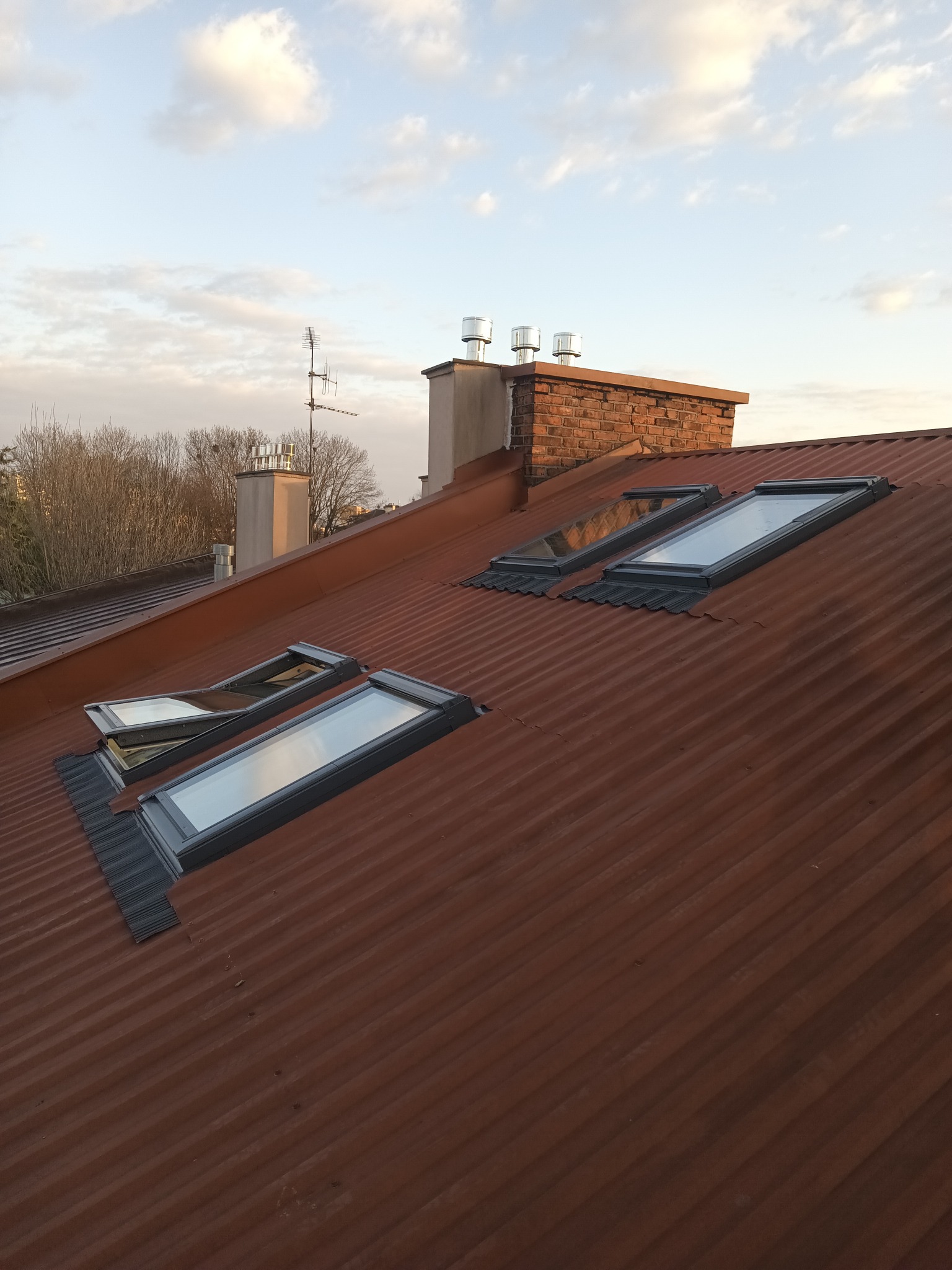 Czerwony, blaszany dach z trzema oknami dachowymi, z których jedno jest otwarte, a w tle widoczny ceglany komin i drzewa.