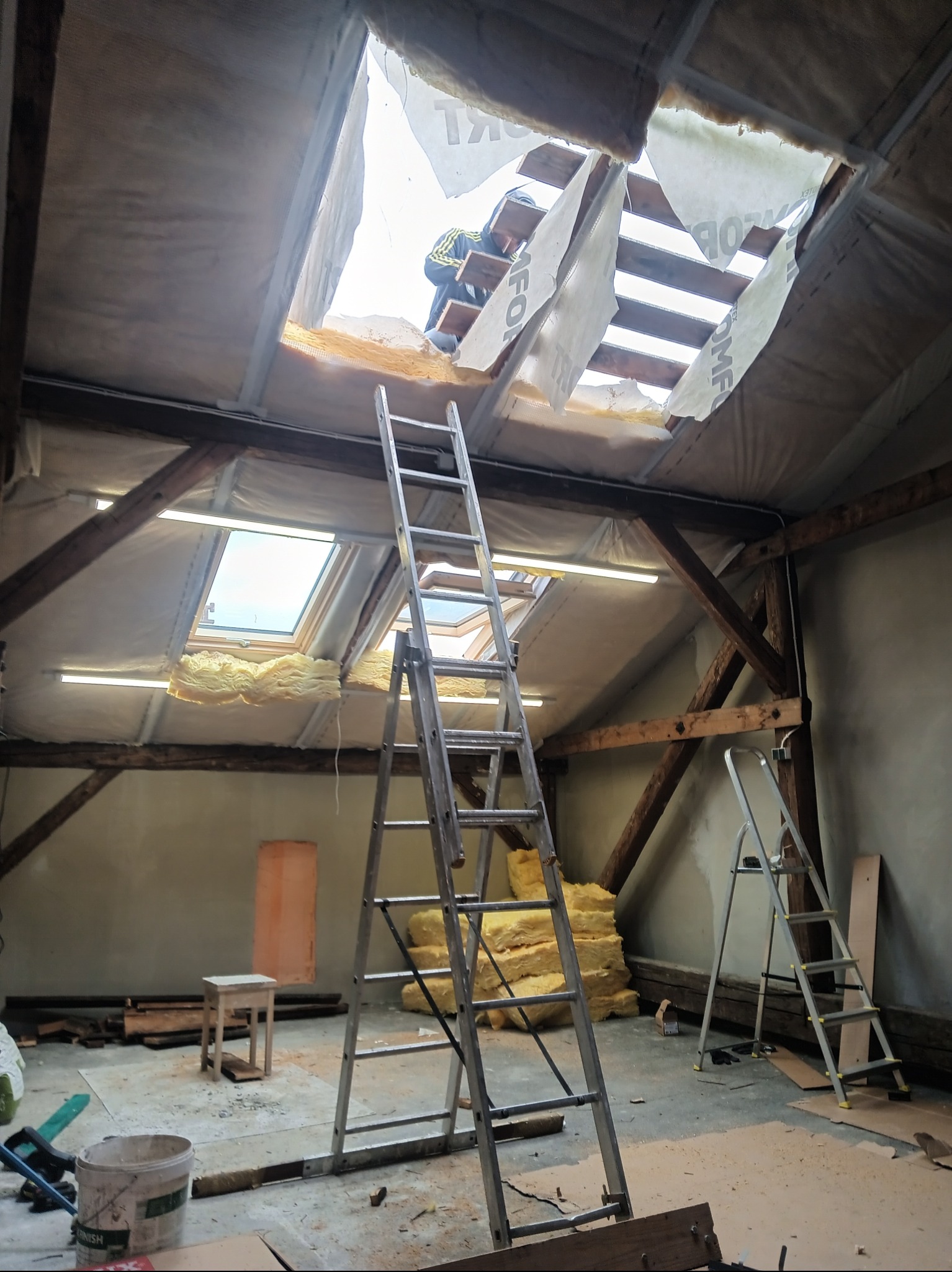 Wnętrze poddasza w trakcie wymiany dachu, widoczne drewniane krokwie, izolacja, folia dachowa z widocznymi napisami, dwie drabiny aluminiowe, stos wełny mineralnej, oraz fragment sylwetki...