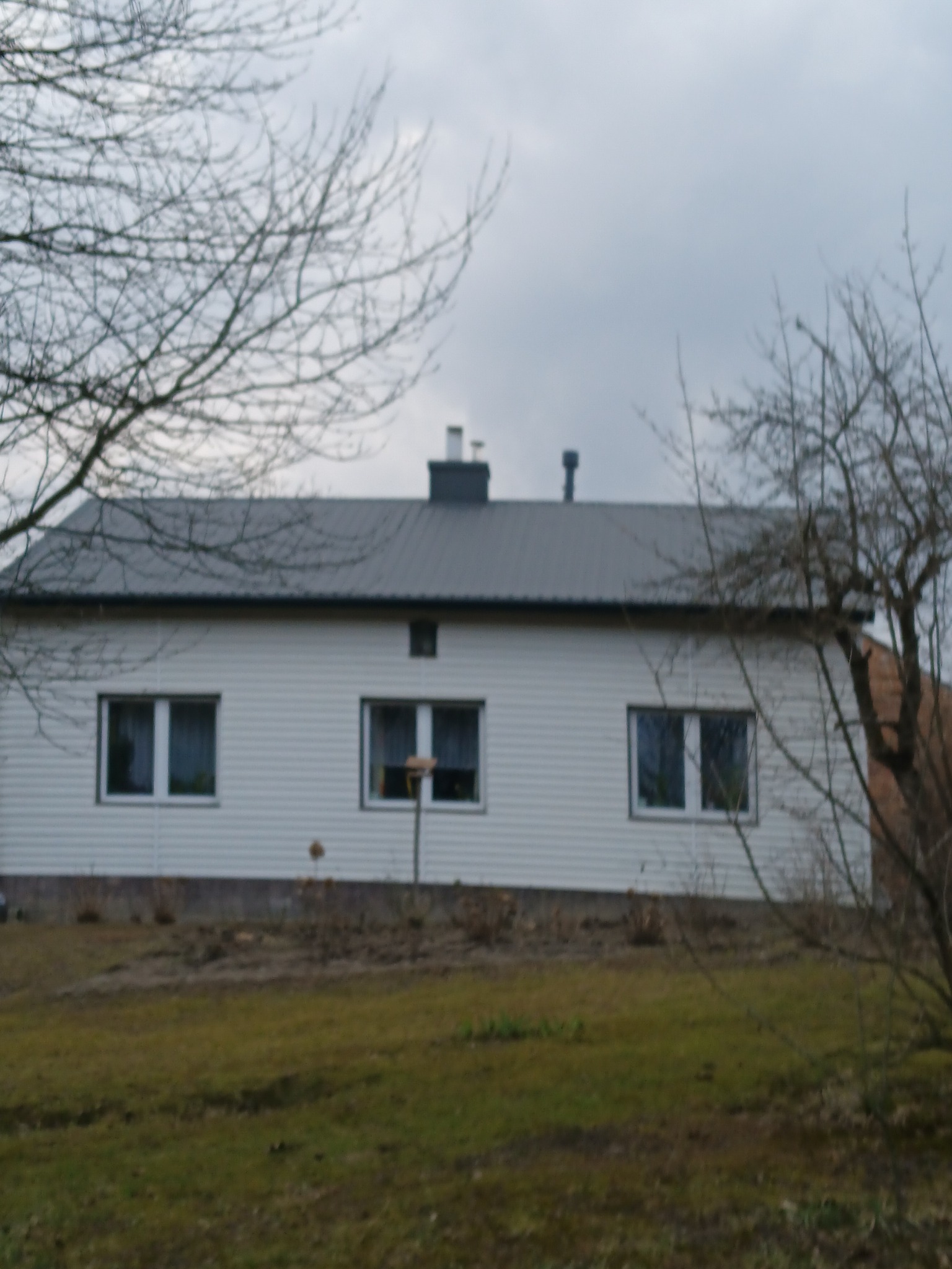 Dom jednorodzinny z szarą, metalową blachą na dachu, widoczny z perspektywy ogrodu w pochmurny dzień.