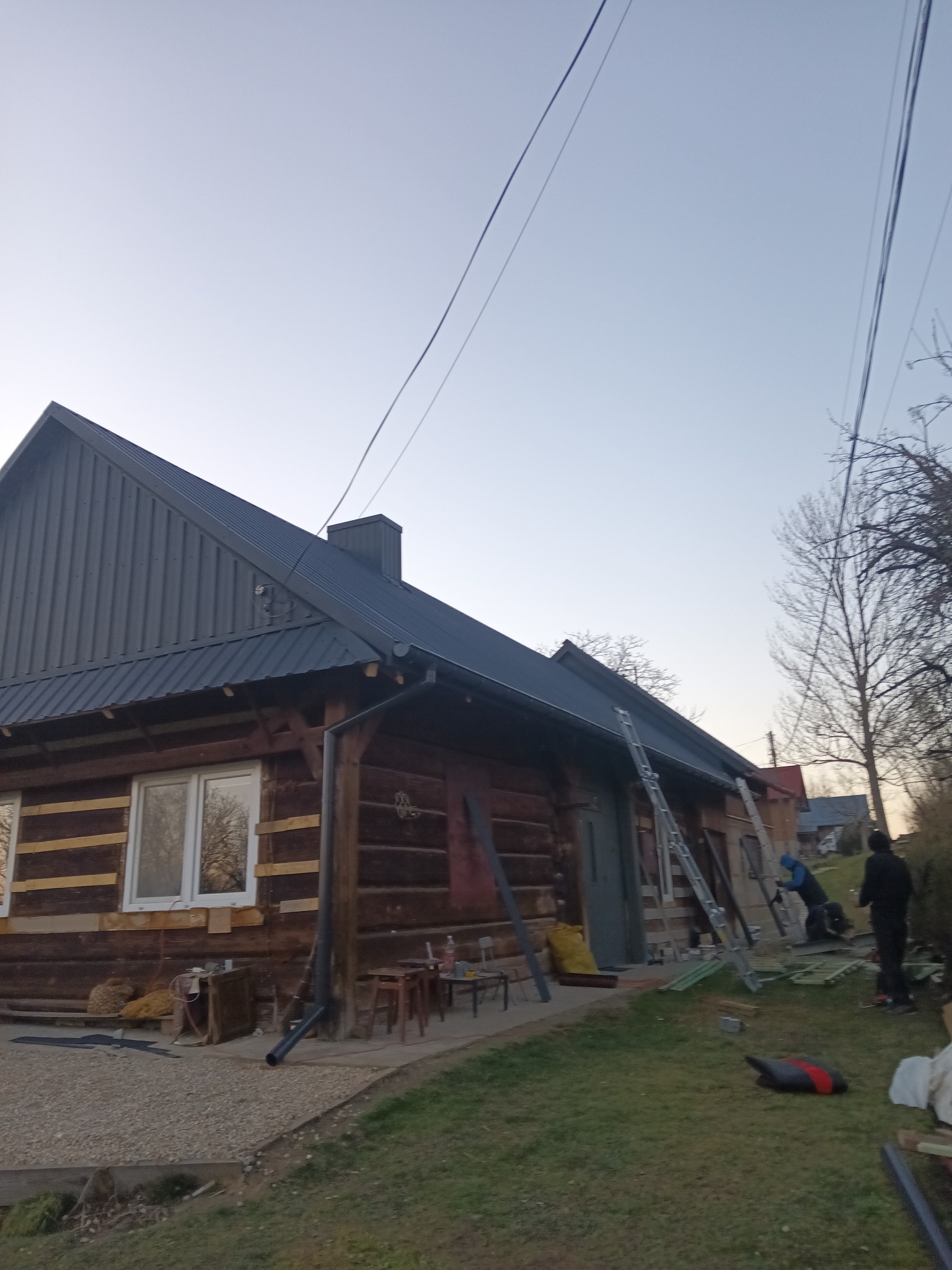Dom z bali z nowym, ciemnoszarym dachem z blachy trapezowej i zamontowanymi rynnami, widoczne drabiny i ekipa montująca.