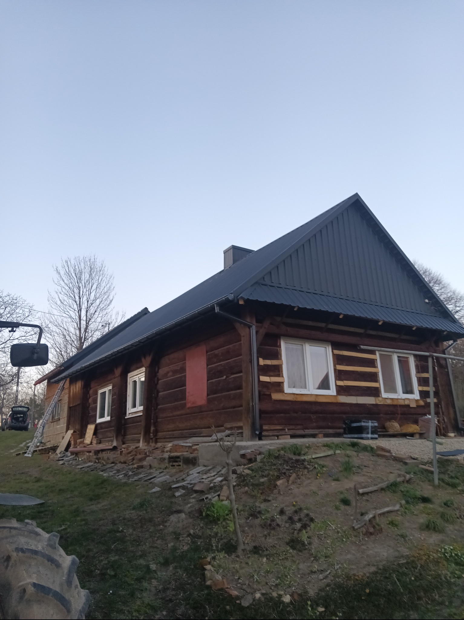 Drewniany dom w trakcie renowacji, z nowym, ciemnym dachem z blachy trapezowej, częściowo ocieplony, widoczne rusztowania i materiały budowlane.