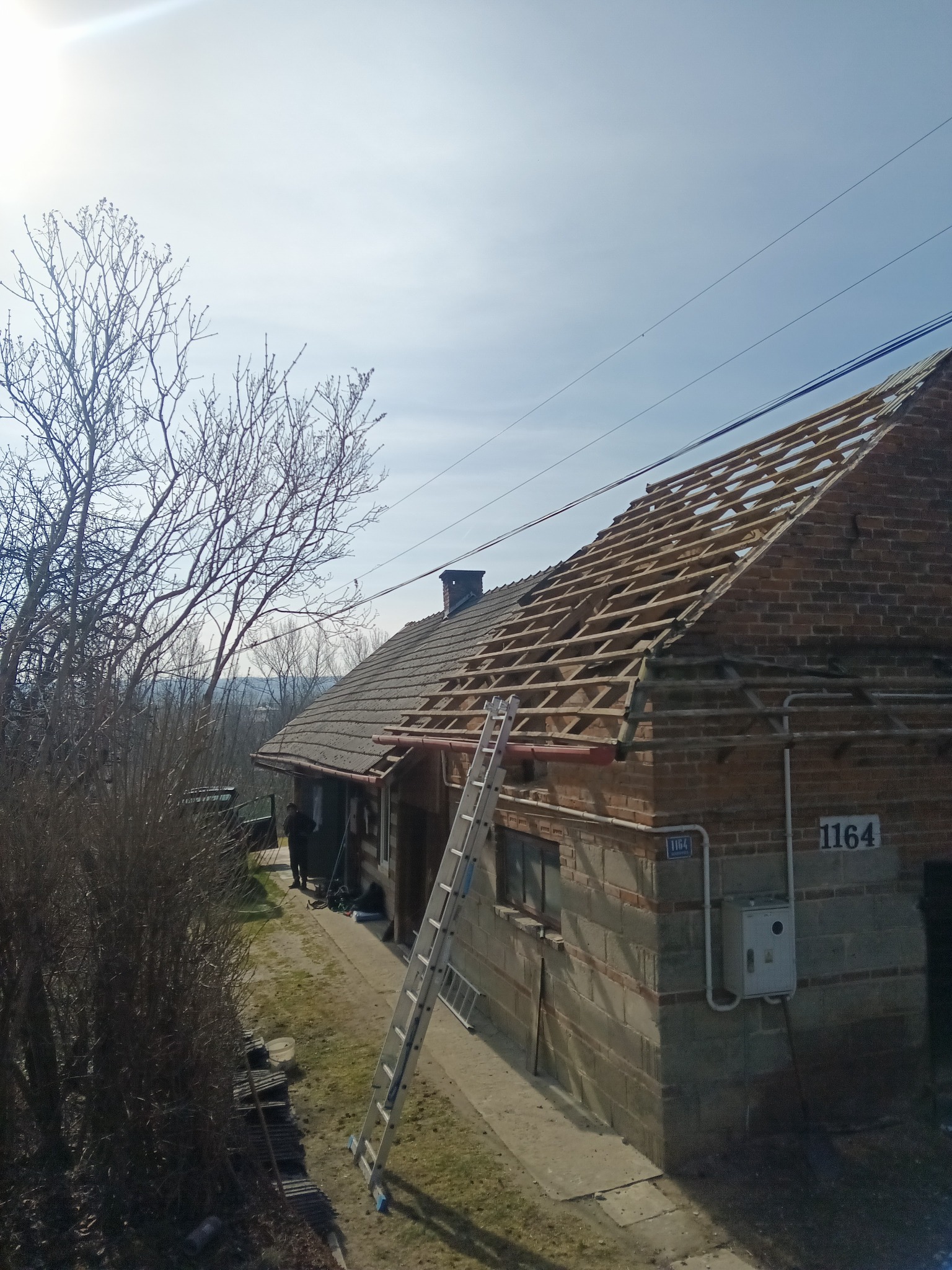 Częściowo zdjęte poszycie dachu z widoczną konstrukcją drewnianą na ceglanym domu jednorodzinnym, drabina oparta o ścianę, w tle drzewa bez liści i błękitne niebo.